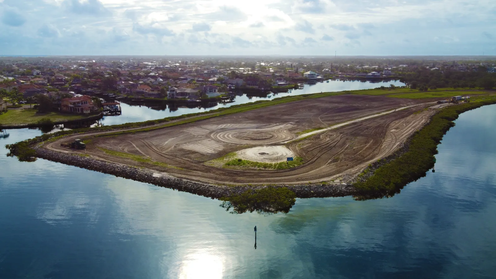 Aerial view of a large cleared land area surrounded by water with residential houses in the background under a cloudy sky.