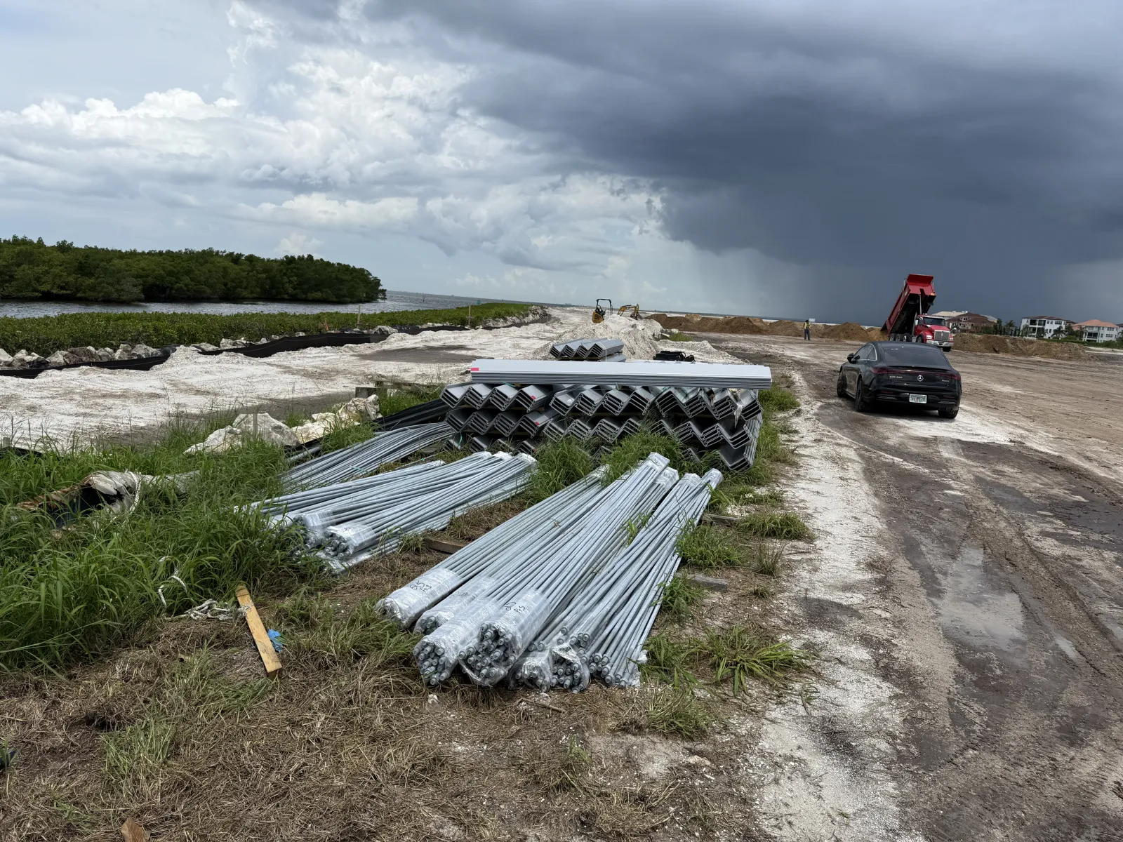 Stacks of metal pipes and construction materials on a dirt road with storm clouds approaching and vehicles nearby