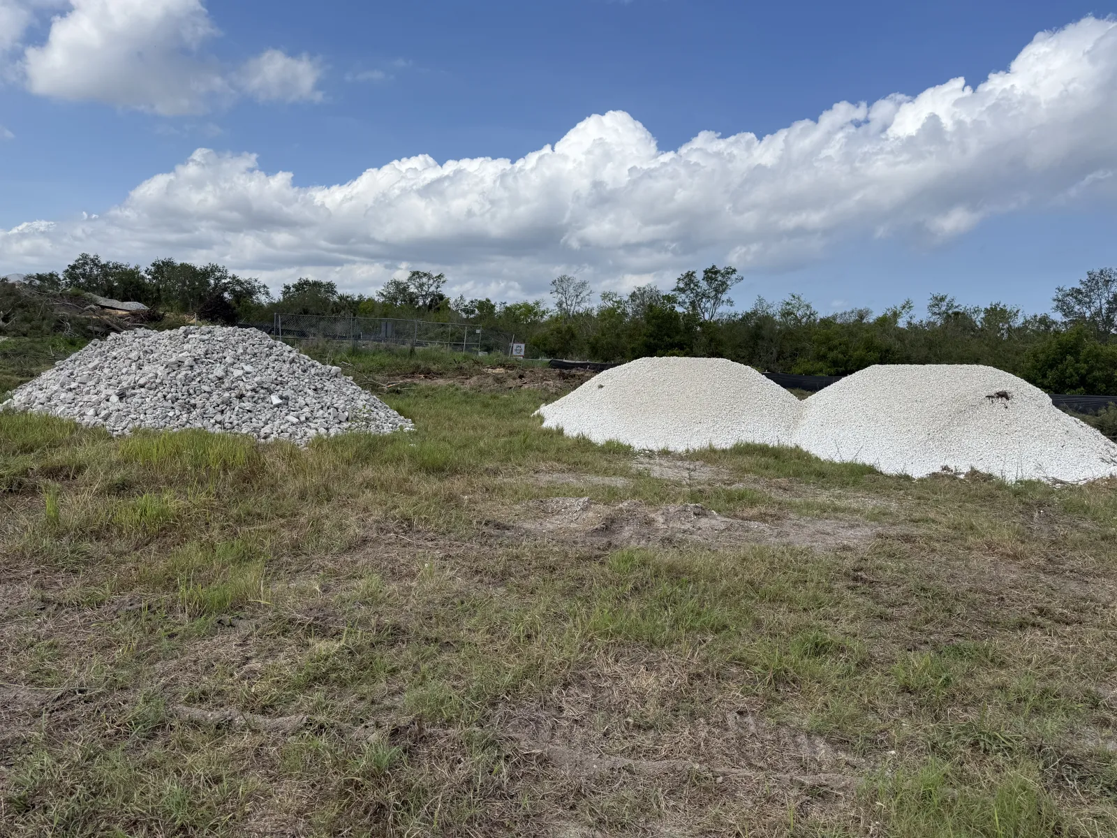 Three piles of gravel and crushed stones on grassy land under a partly cloudy blue sky near trees.
