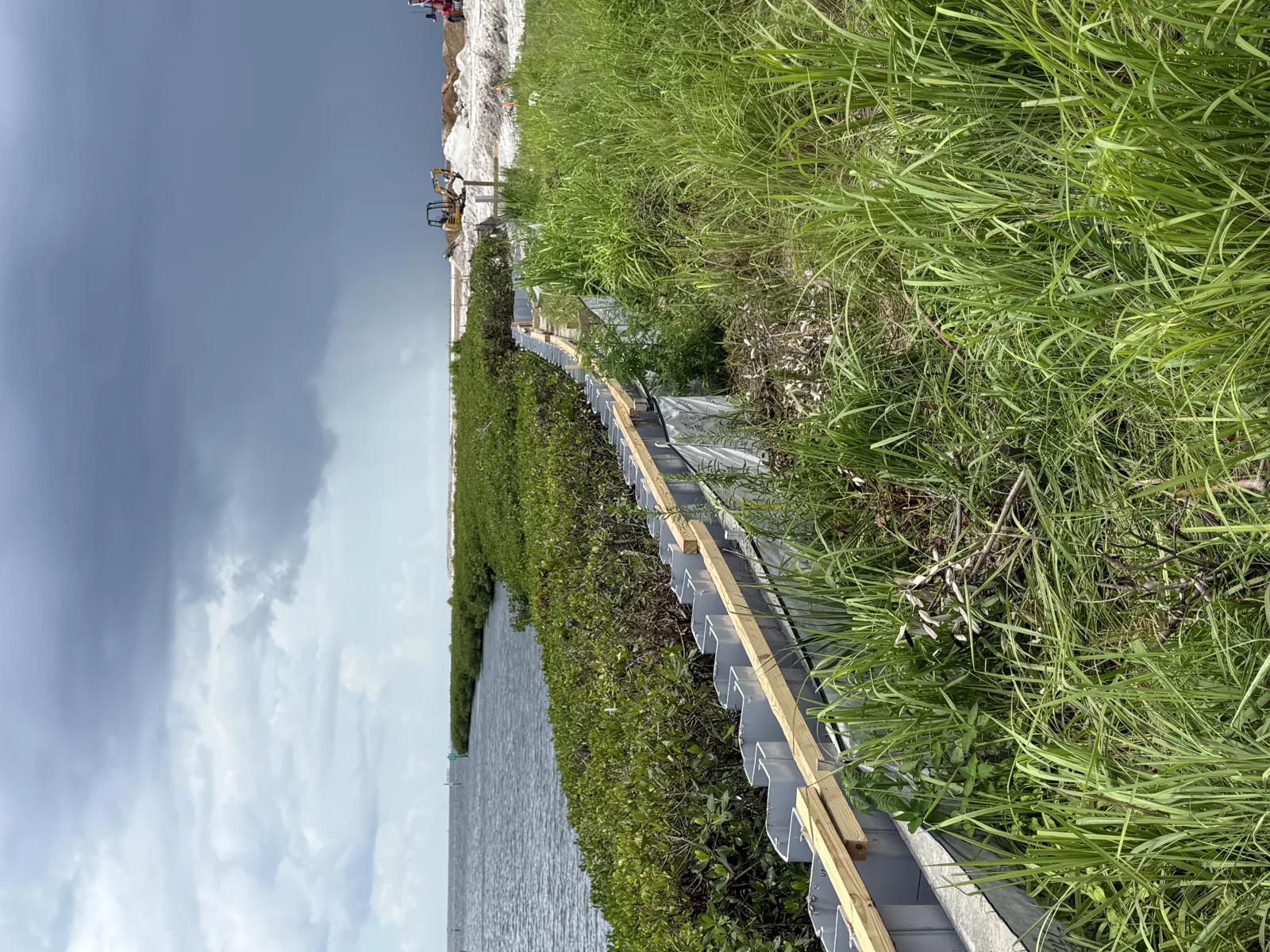 Waterway bordered by vegetation and grass under a cloudy sky with a wooden structure along the shore.