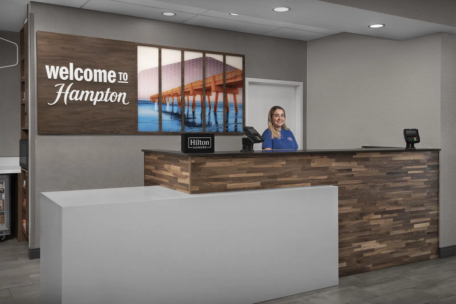 Reception desk at Hampton hotel with a friendly staff member and welcome sign featuring a pier over water.