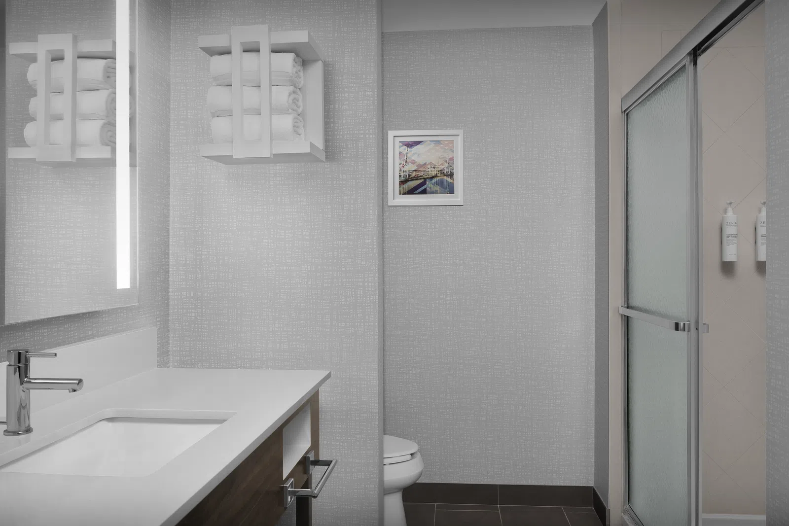 Modern bathroom with white countertop, wooden vanity, glass shower door, and towels on wall shelves.