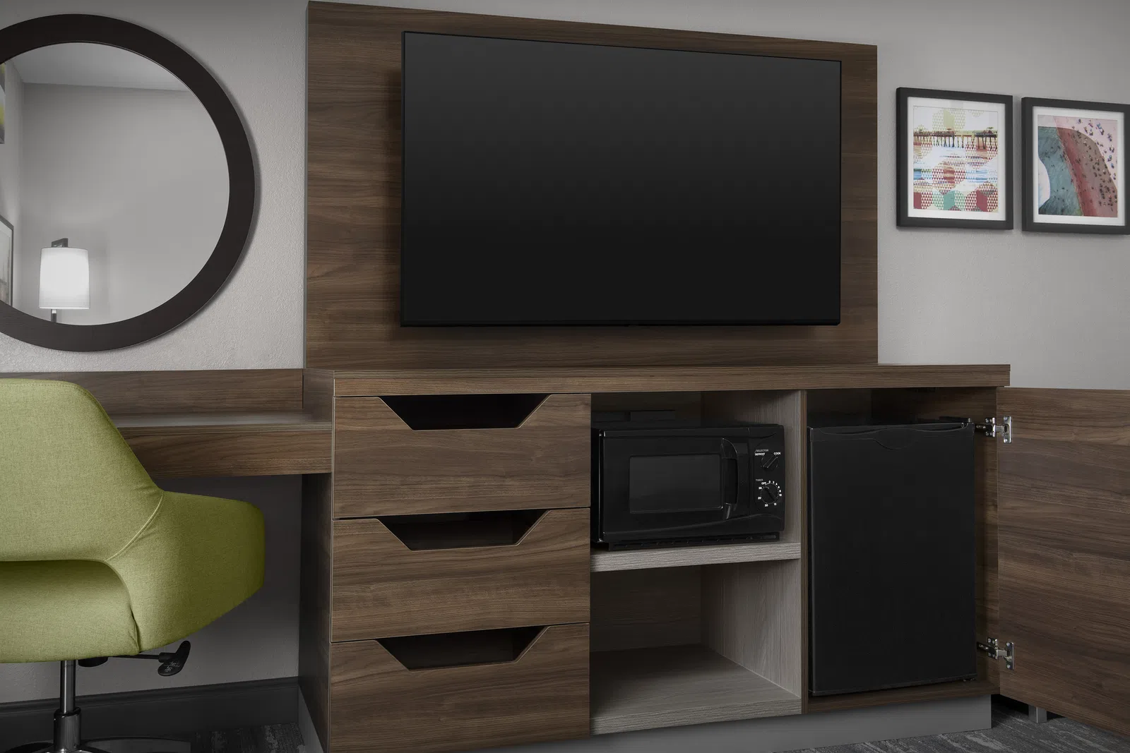 Modern hotel room corner with wood TV stand, mounted flat-screen TV, microwave, mini fridge, green chair, and wall art.