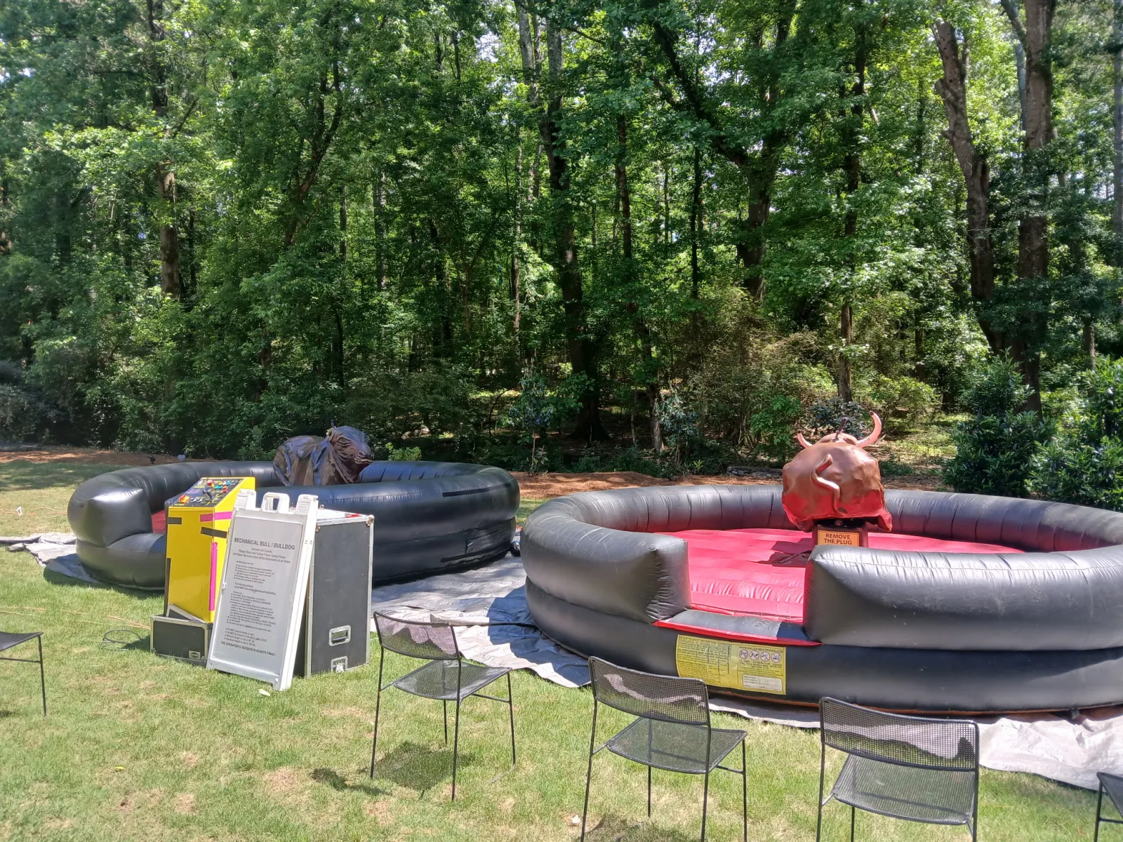 Outdoor mechanical bull riding setup with two inflatable arenas on grass surrounded by chairs and trees.