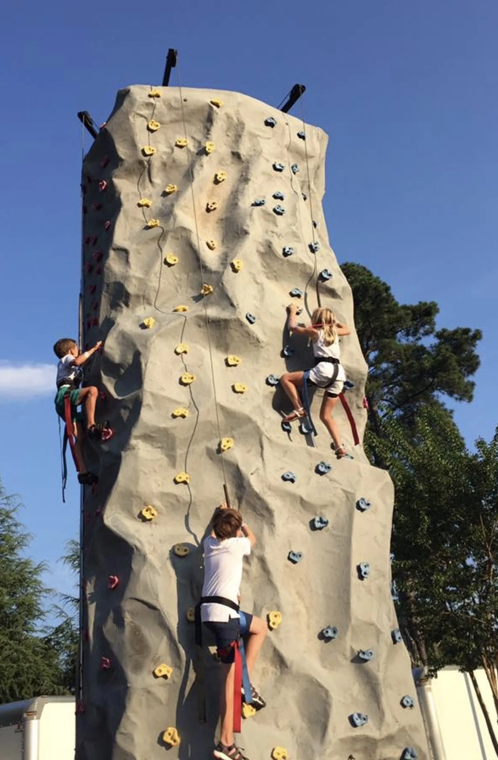 Three children climbing a large outdoor rock climbing wall with safety harnesses on a clear sunny day.