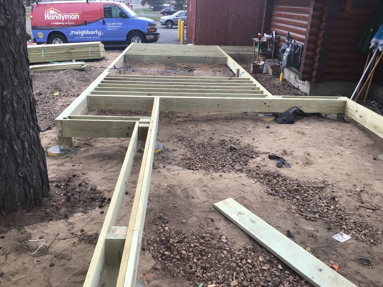 Wooden frame structure for a deck or small building under construction on dirt ground near a log cabin.