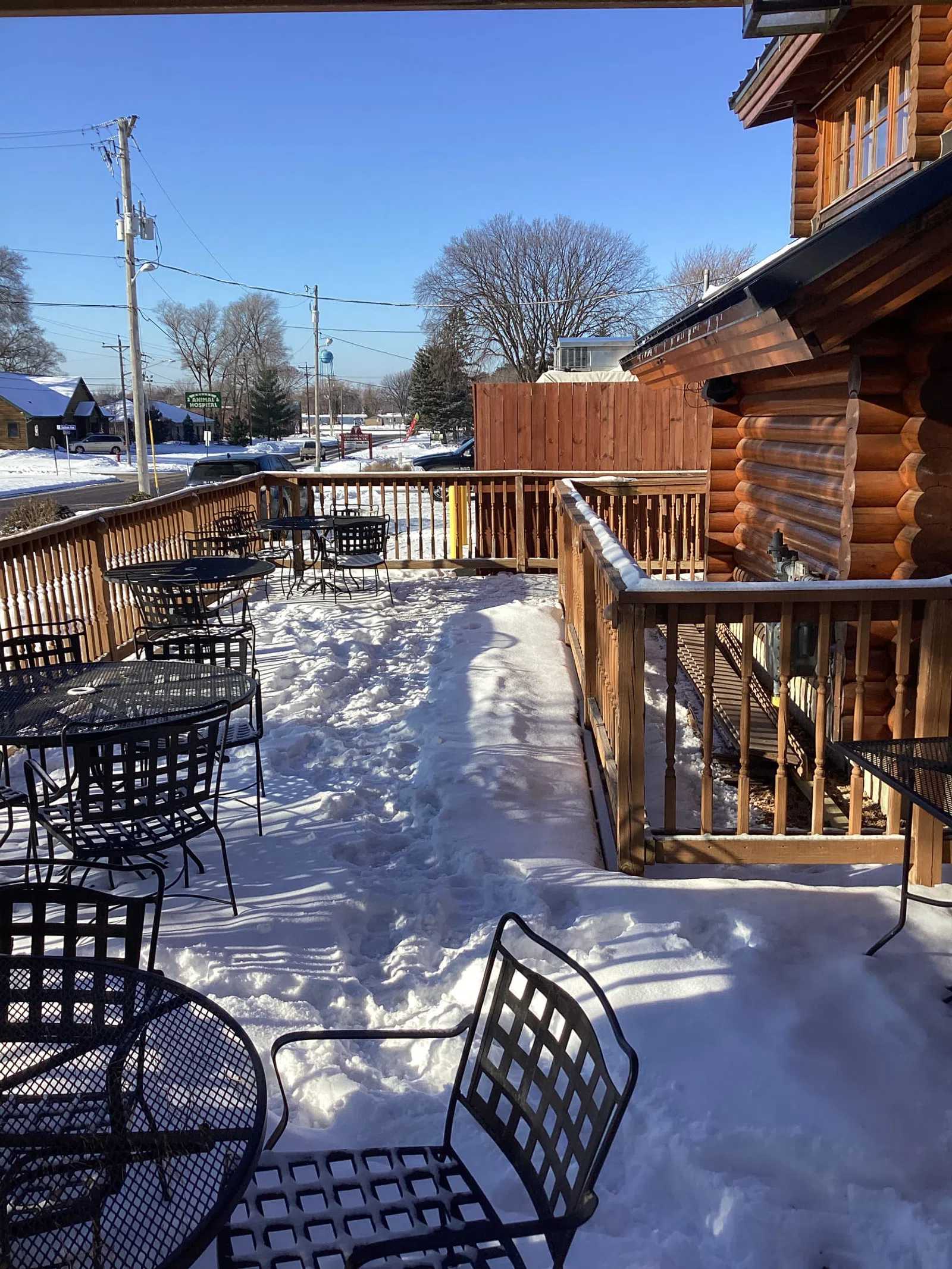 Snow-covered outdoor patio with metal tables and chairs next to a wooden log cabin under clear blue sky.