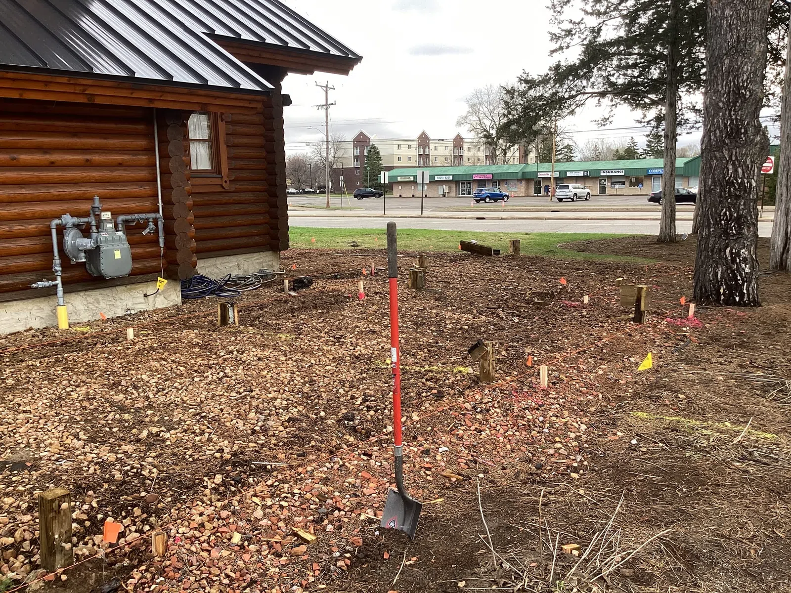 Shovel standing upright in prepared soil beside a wooden log cabin with commercial buildings in background