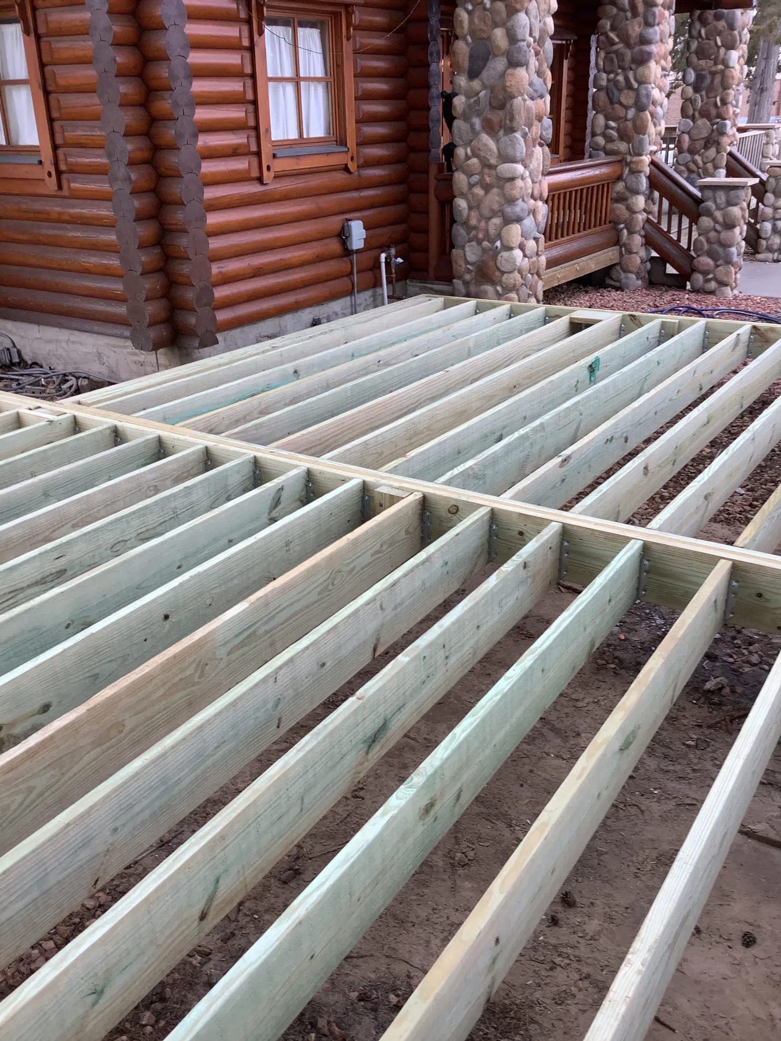 Wooden deck frame under construction next to a log cabin with stone porch columns outdoors.
