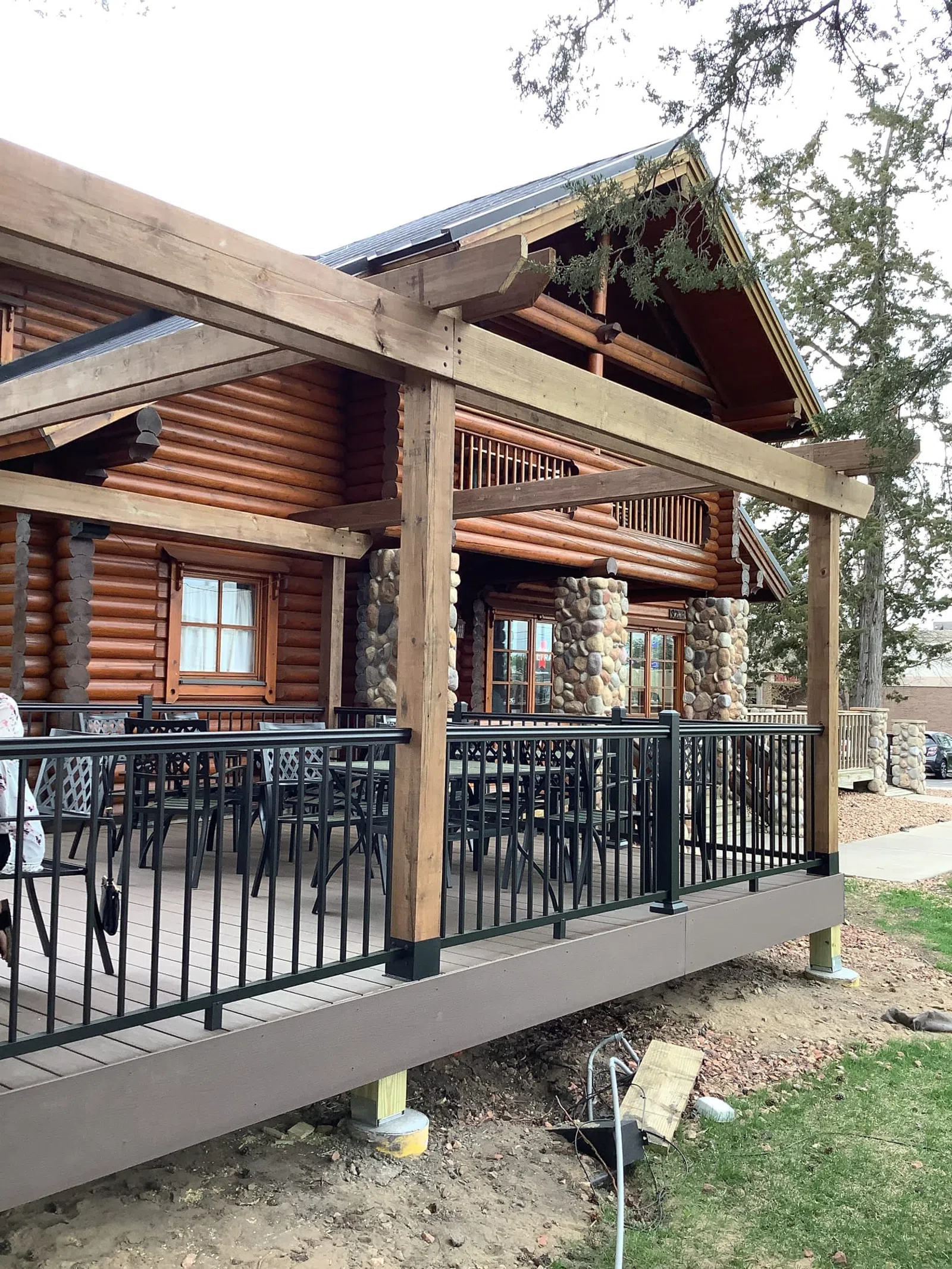 Log cabin porch with wooden beams, stone pillars, black metal railing, and outdoor seating under trees.