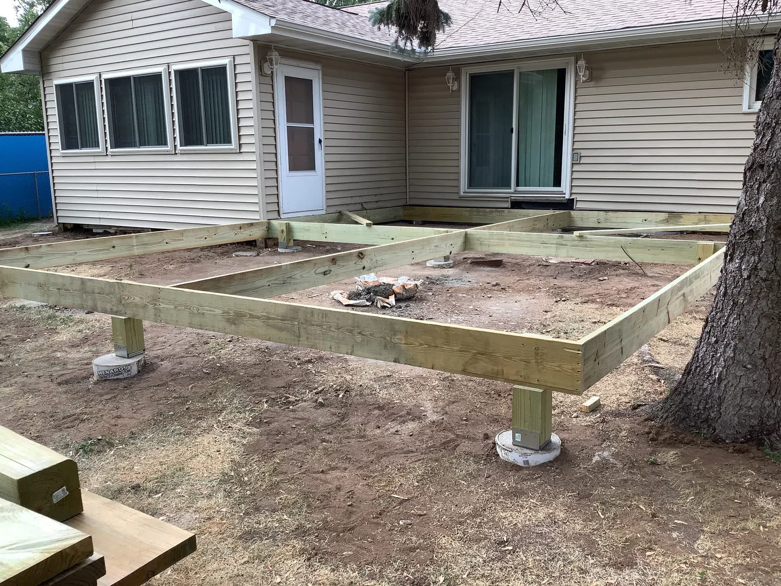 Wooden deck frame construction supported by concrete piers next to a beige house with windows and sliding door.