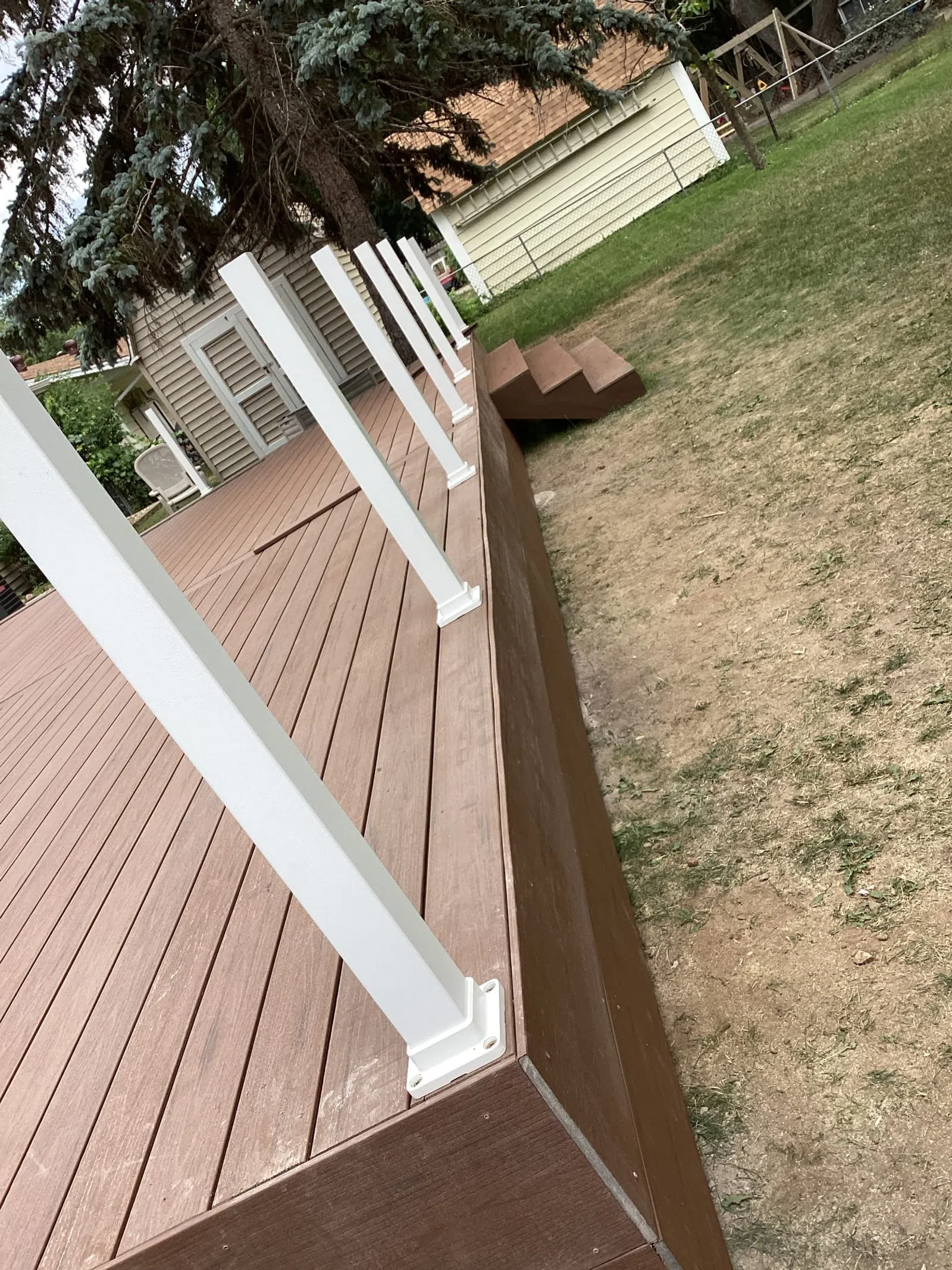 New brown wooden deck with white railing posts and small stairs leading to a backyard with patchy grass.