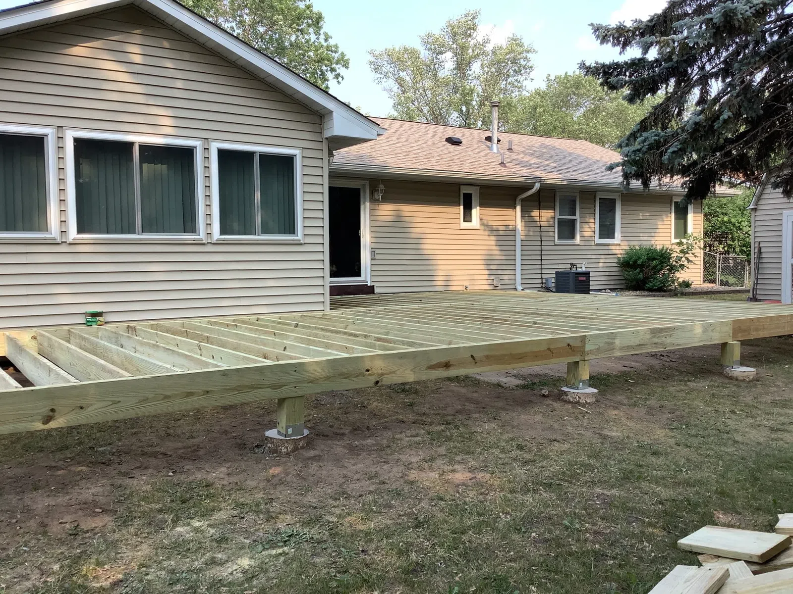 Wooden deck framework under construction beside a single-story house in a backyard with trees and grass.