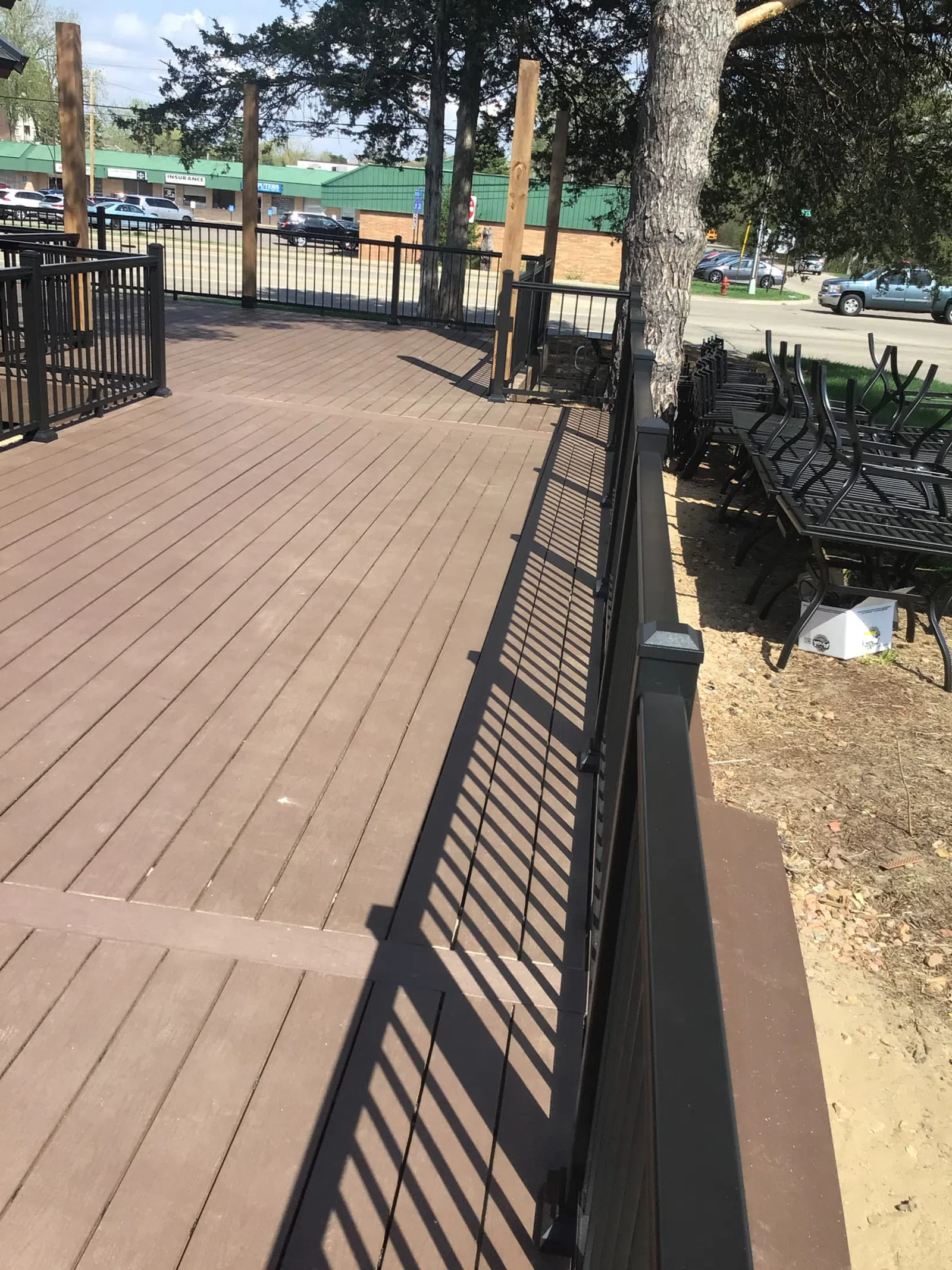 Empty brown wooden deck with black metal railing and stacked outdoor chairs beside trees in sunny weather.