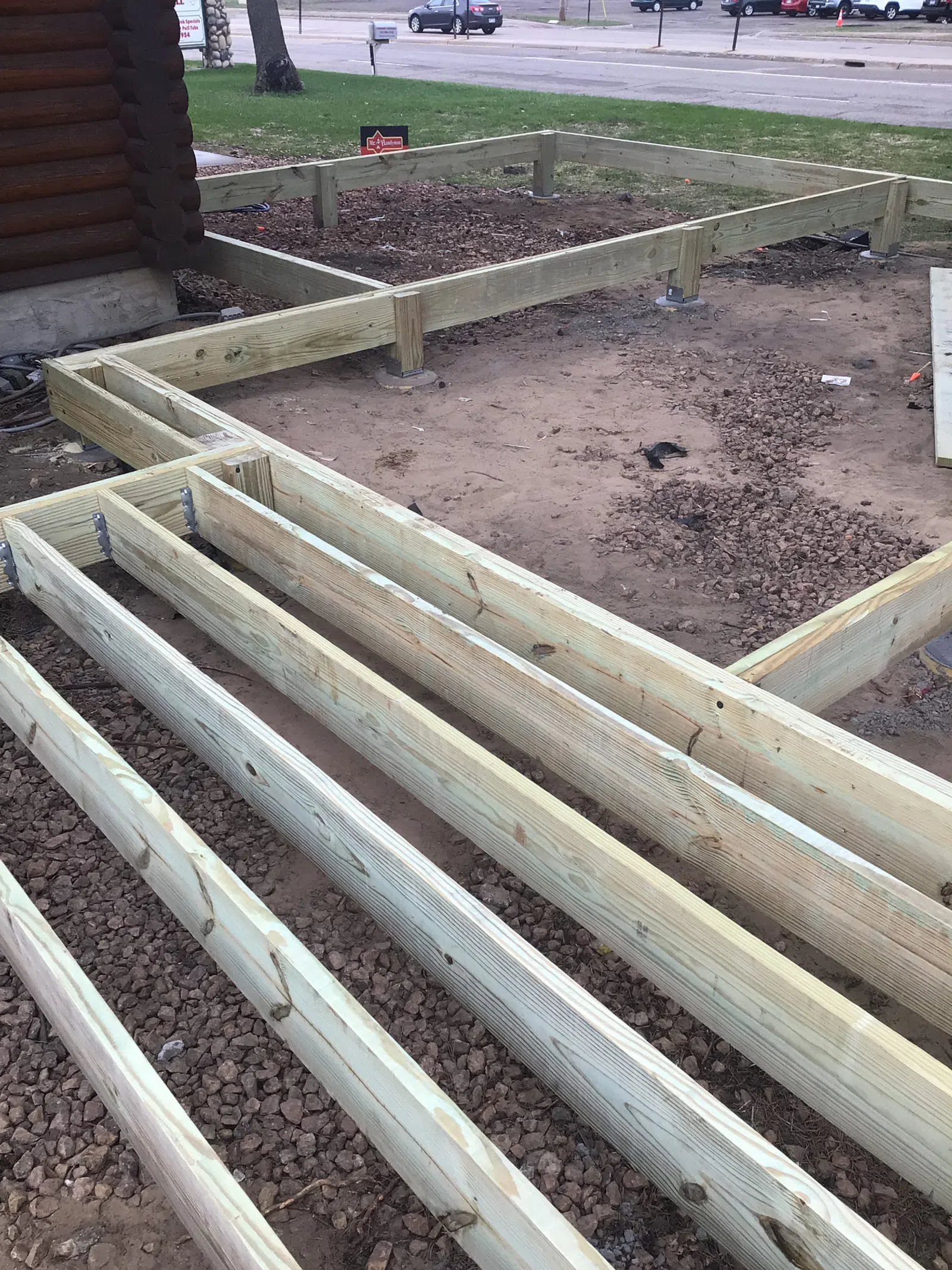 Wooden frame foundation construction for a deck or small building next to a log cabin on gravel and dirt.