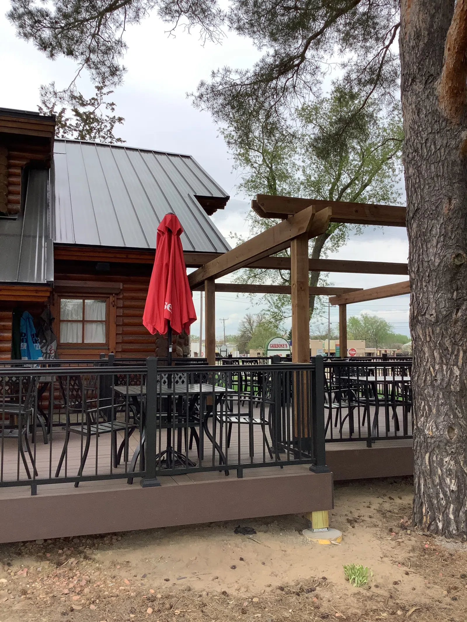 Outdoor seating area with black metal chairs and tables, red closed umbrella, wooden pergola, and log cabin exterior.