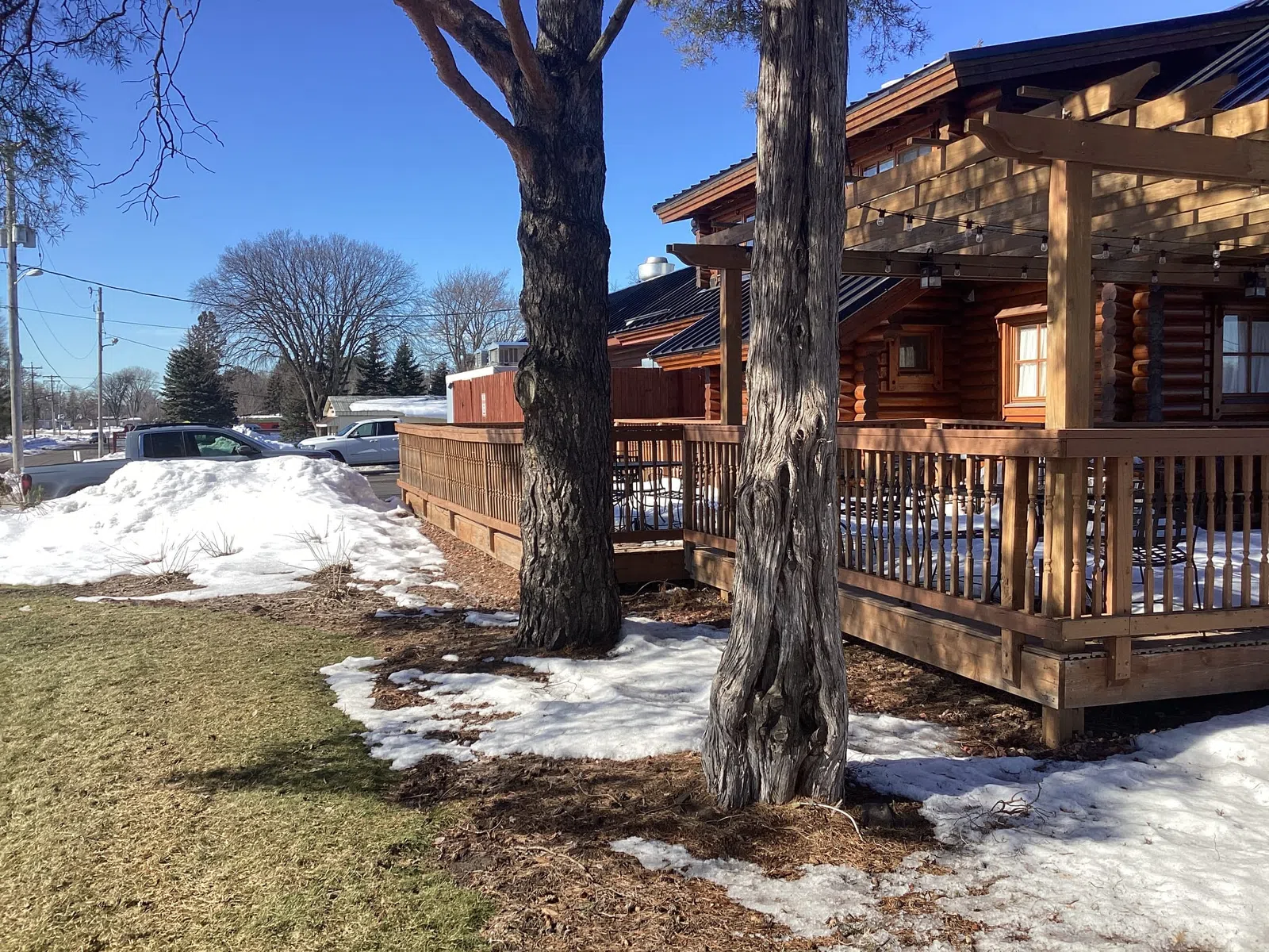 Log cabin with wooden deck surrounded by melting snow and two large trees on a clear sunny day.