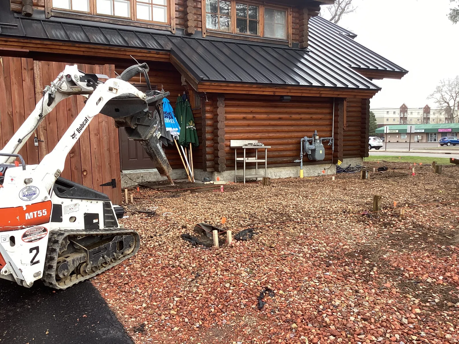 Mini excavator with auger attachment clearing rocky soil beside wooden cabin building under construction.