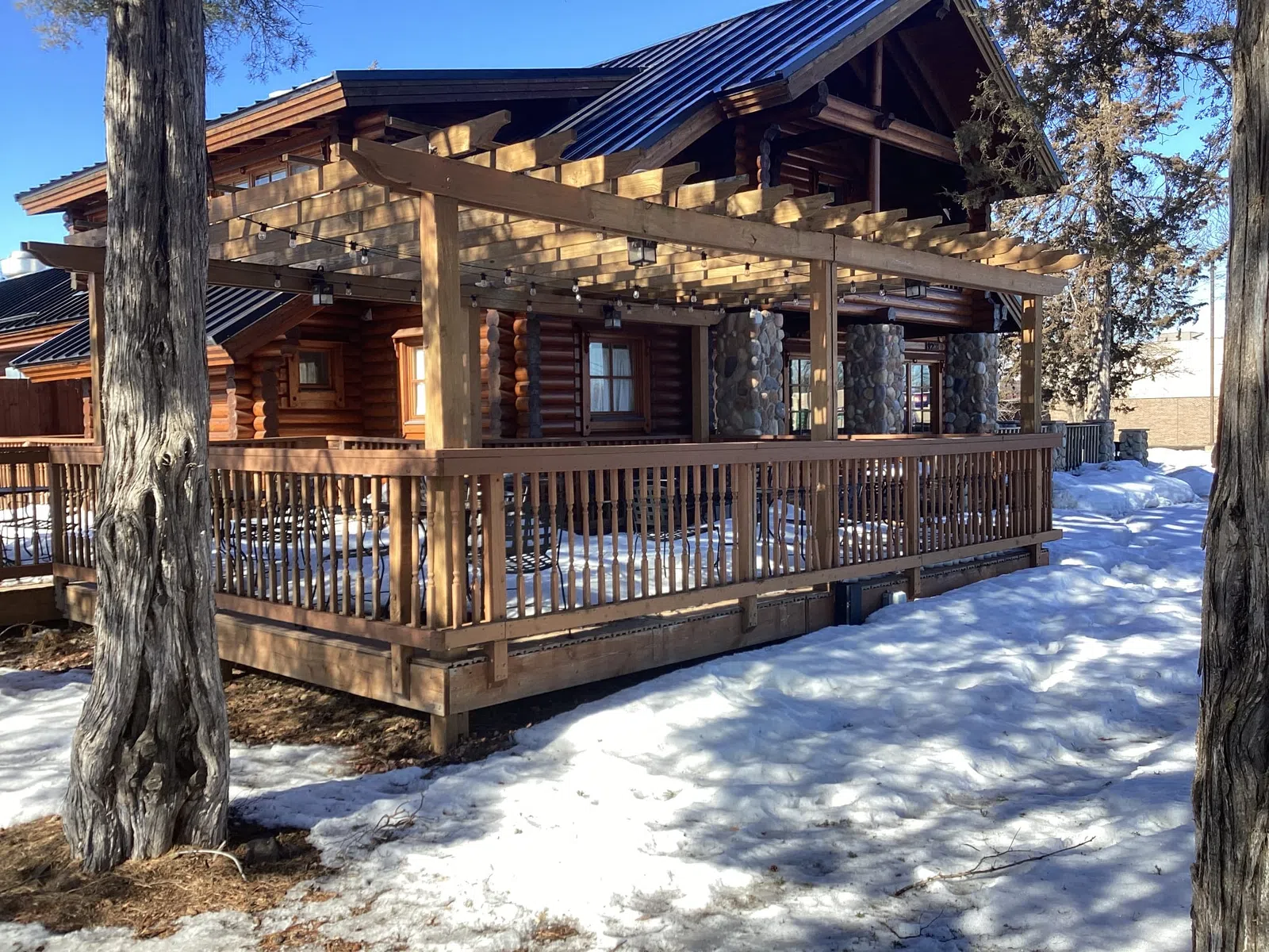 Wooden log cabin with stone chimney surrounded by snow and tall trees under clear blue sky.