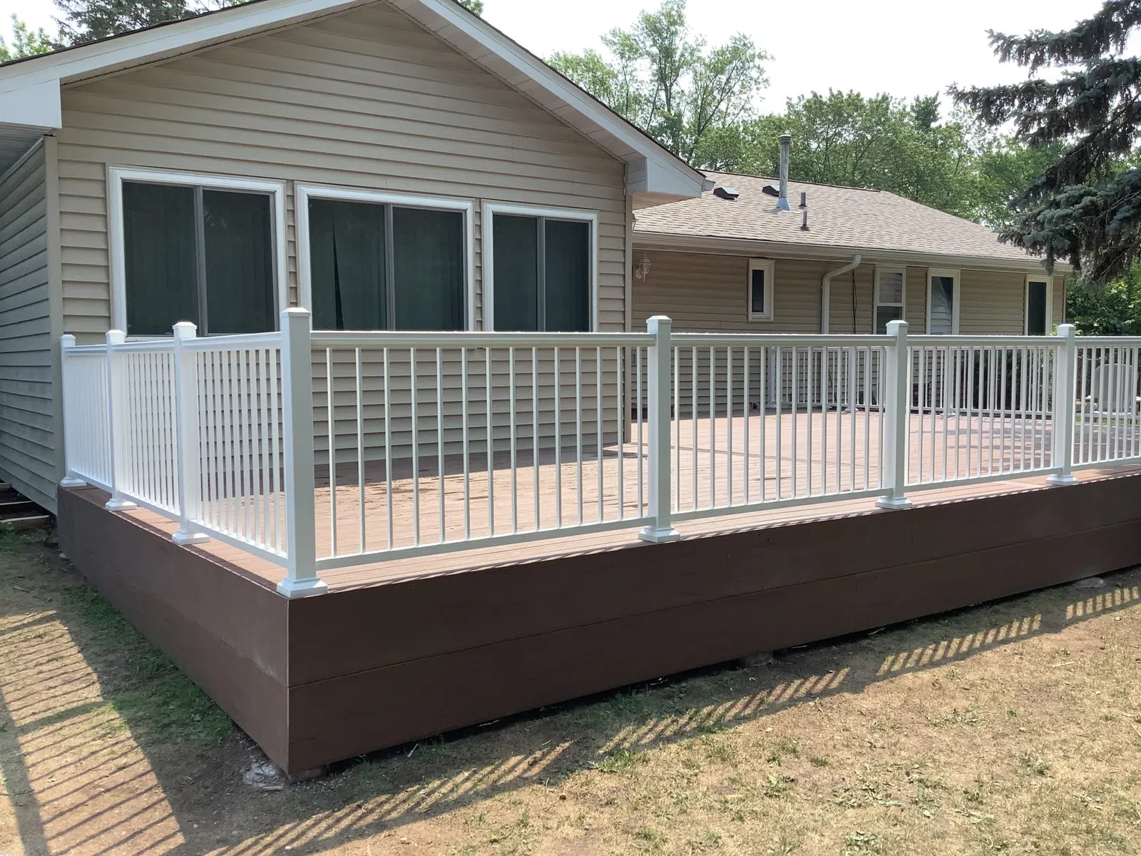 Spacious elevated wooden deck with white railing attached to beige house surrounded by greenery on a sunny day