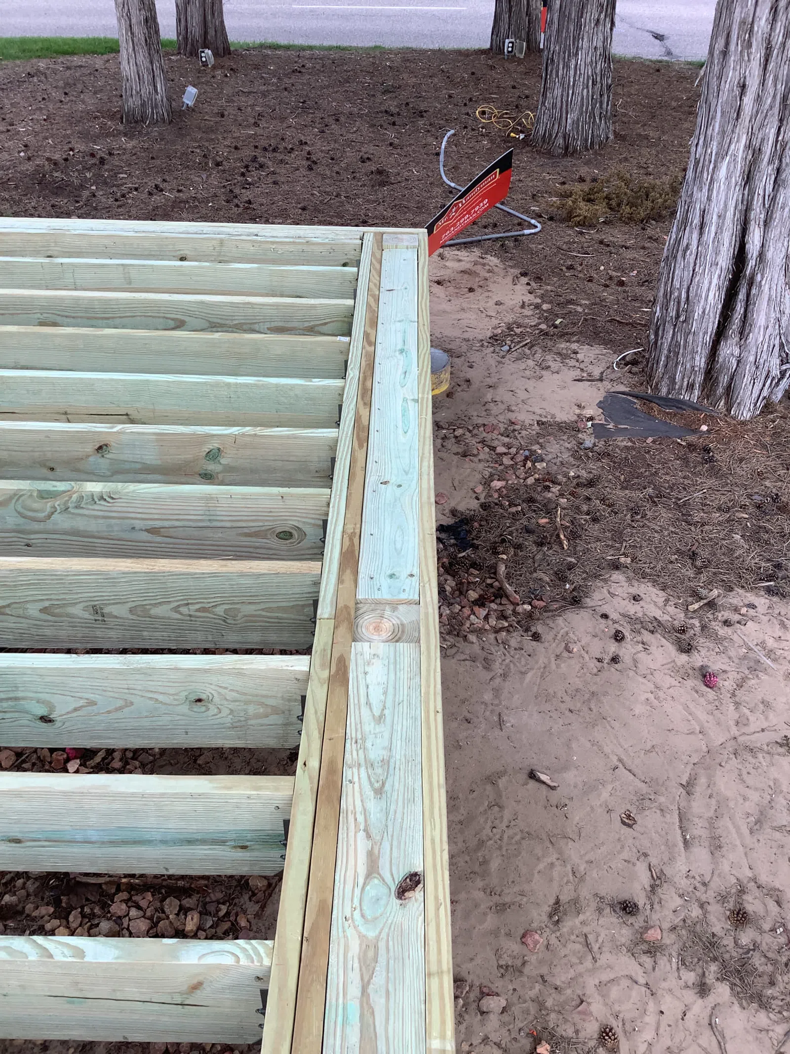 Close-up of wooden deck framing with beams and joists under construction near tree trunks and dirt ground.