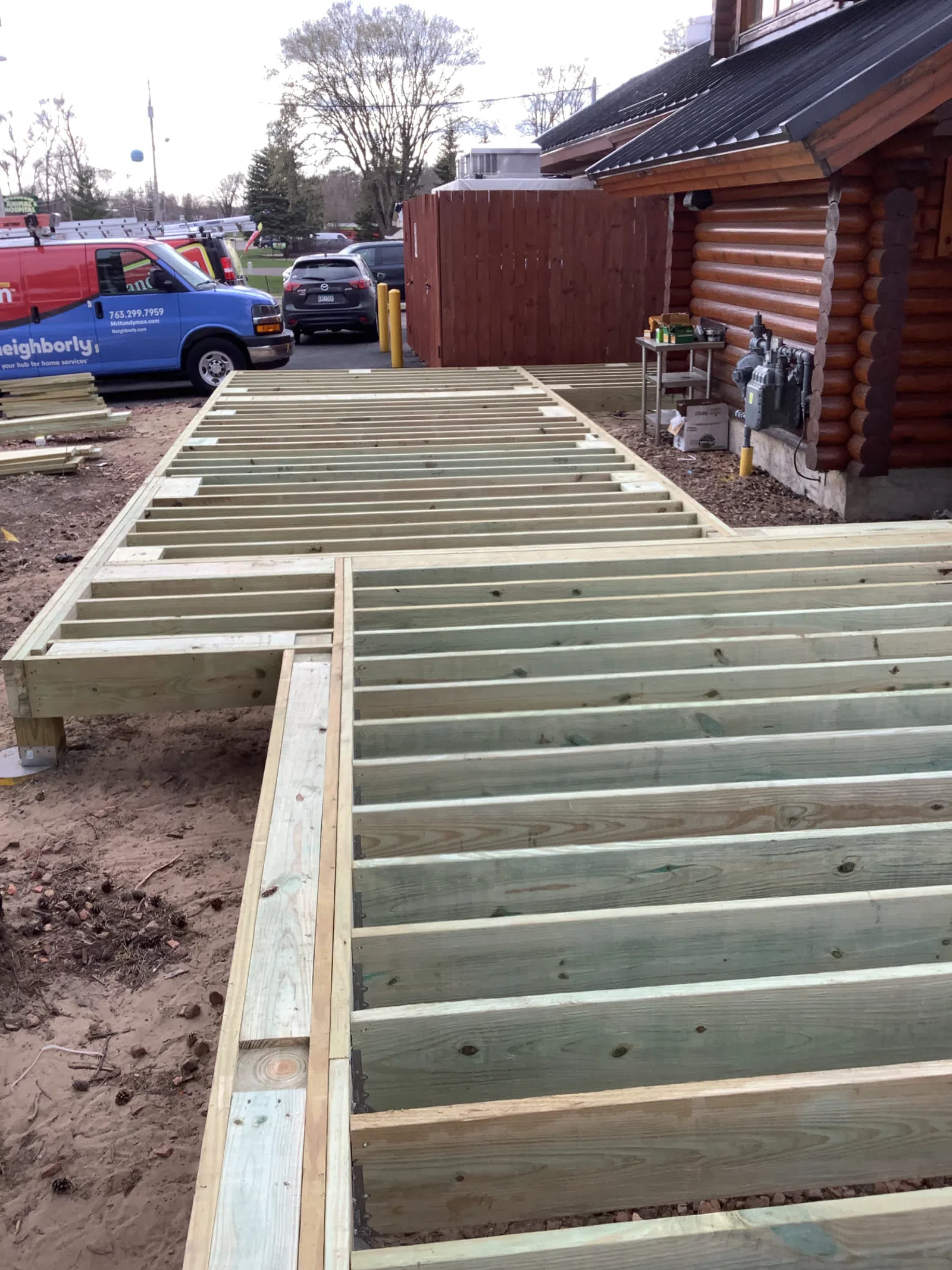 Wooden deck frame construction beside a log cabin on a residential property with work vans nearby