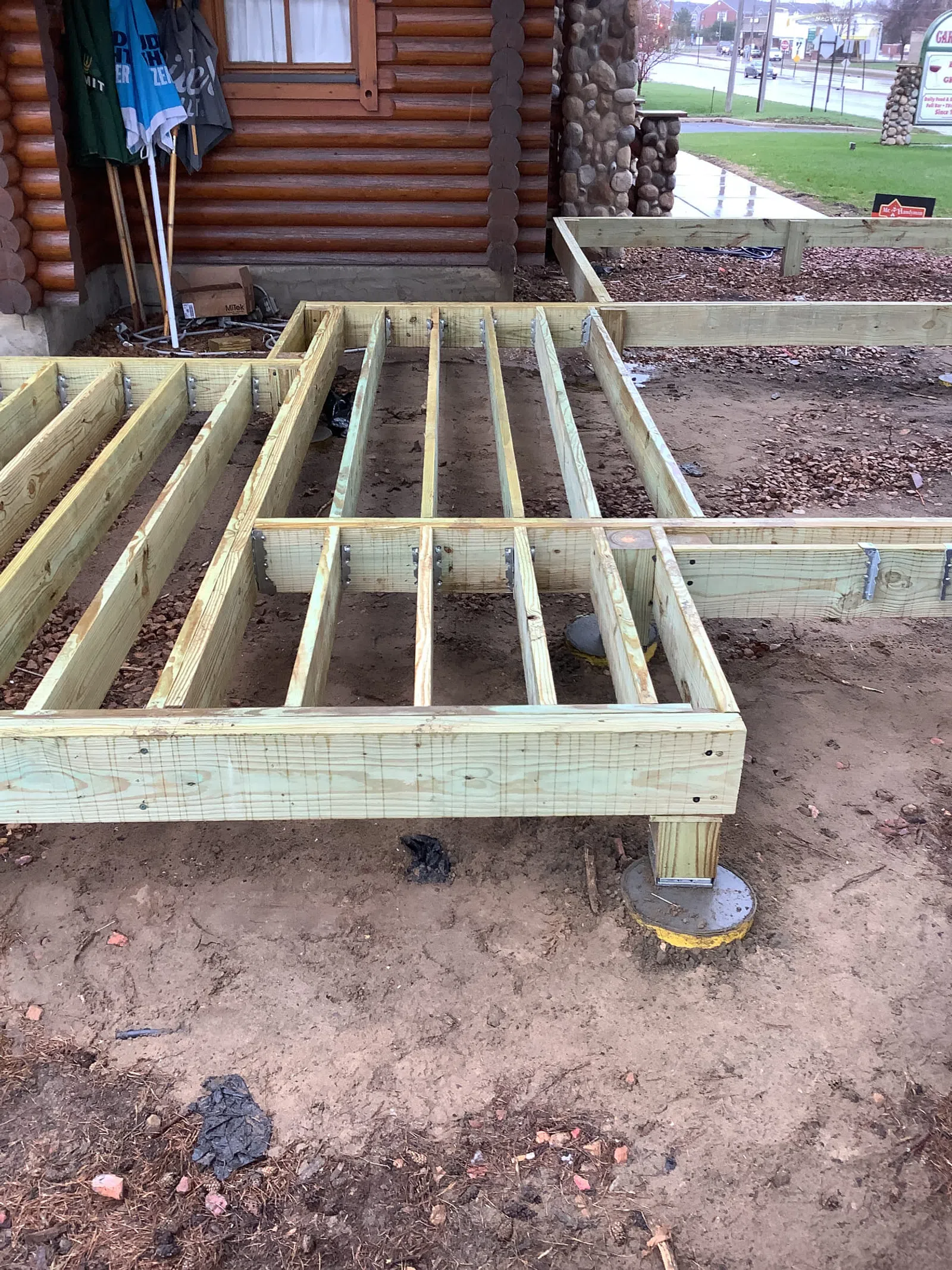 Wooden deck frame construction with metal supports next to a log cabin on a dirt ground.