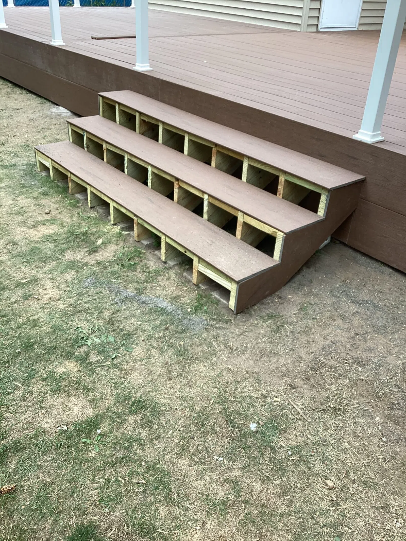Three-step brown wooden outdoor stairs attached to a deck with visible support structure on dry lawn.