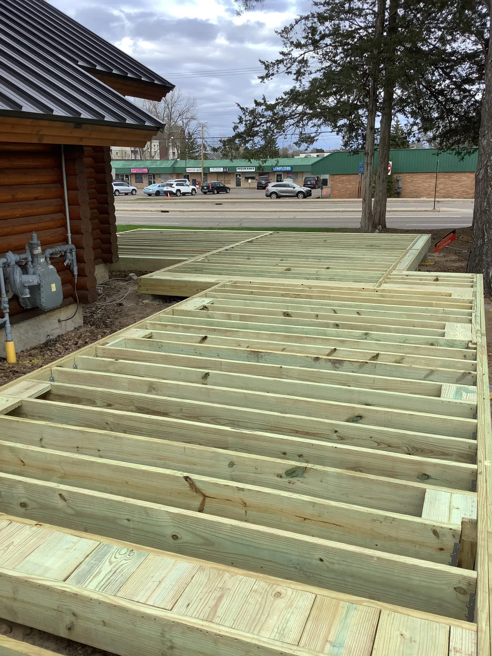 Close-up of wooden deck framing attached to log cabin exterior under cloudy sky with street and buildings in background