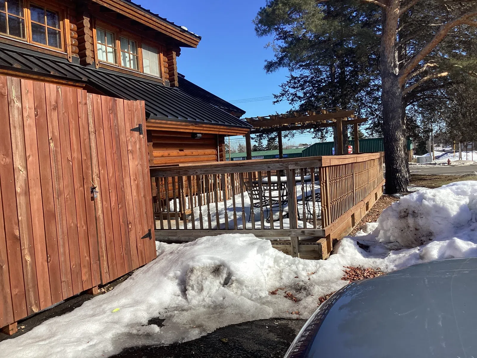 Snow-covered ground around a wooden cabin with a porch railing and clear blue sky on a sunny day.