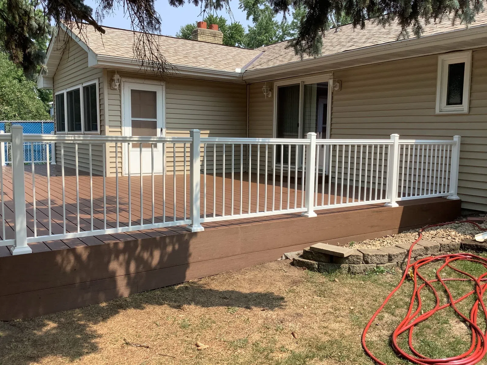 Raised brown wooden deck with white railings attached to beige house under tree shade in backyard.