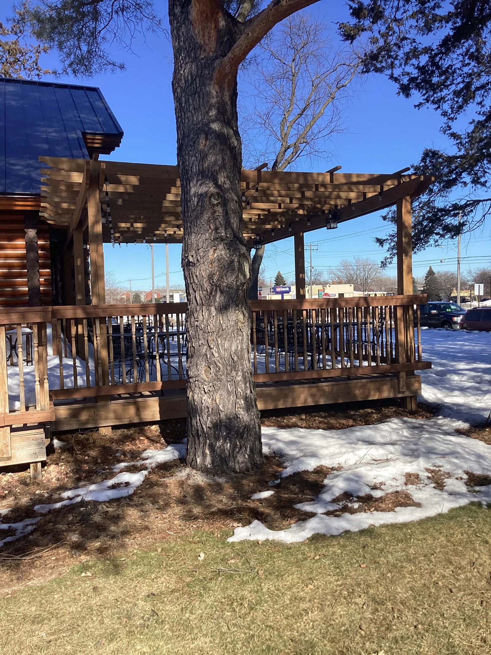 Wooden pergola and deck around a tree with patches of snow and dry grass under a clear blue sky.