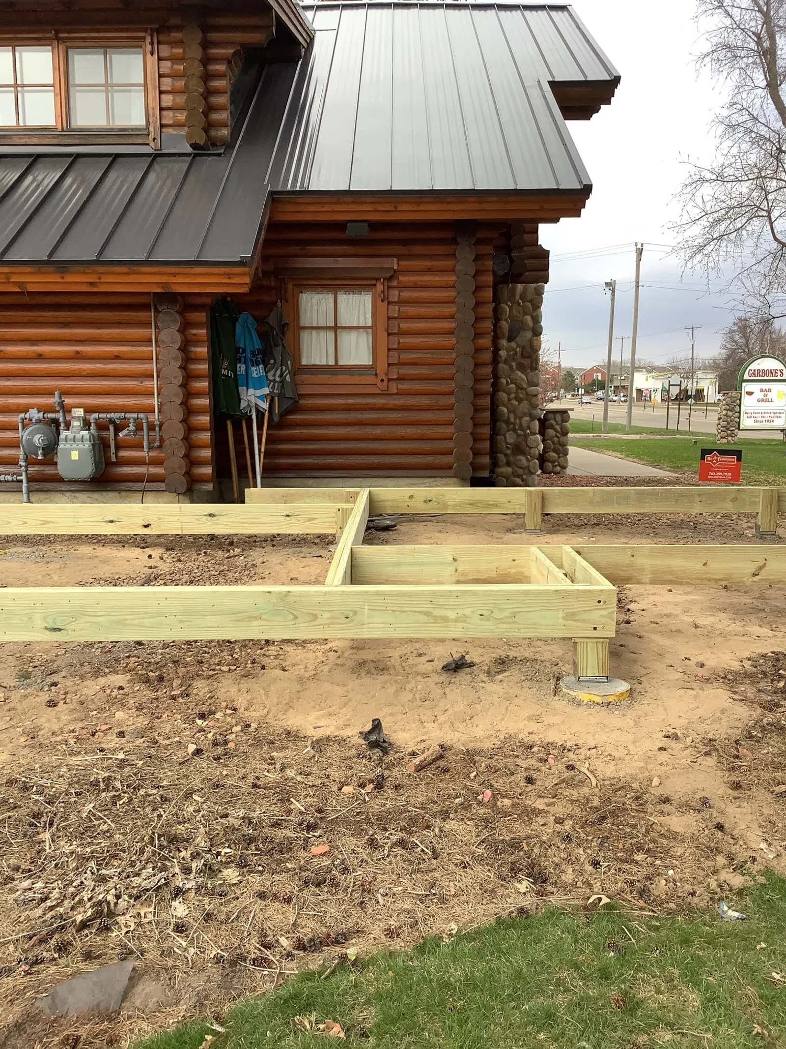 Wooden frame construction beside a log cabin with stone accents and metal roof in an outdoor setting near a street.