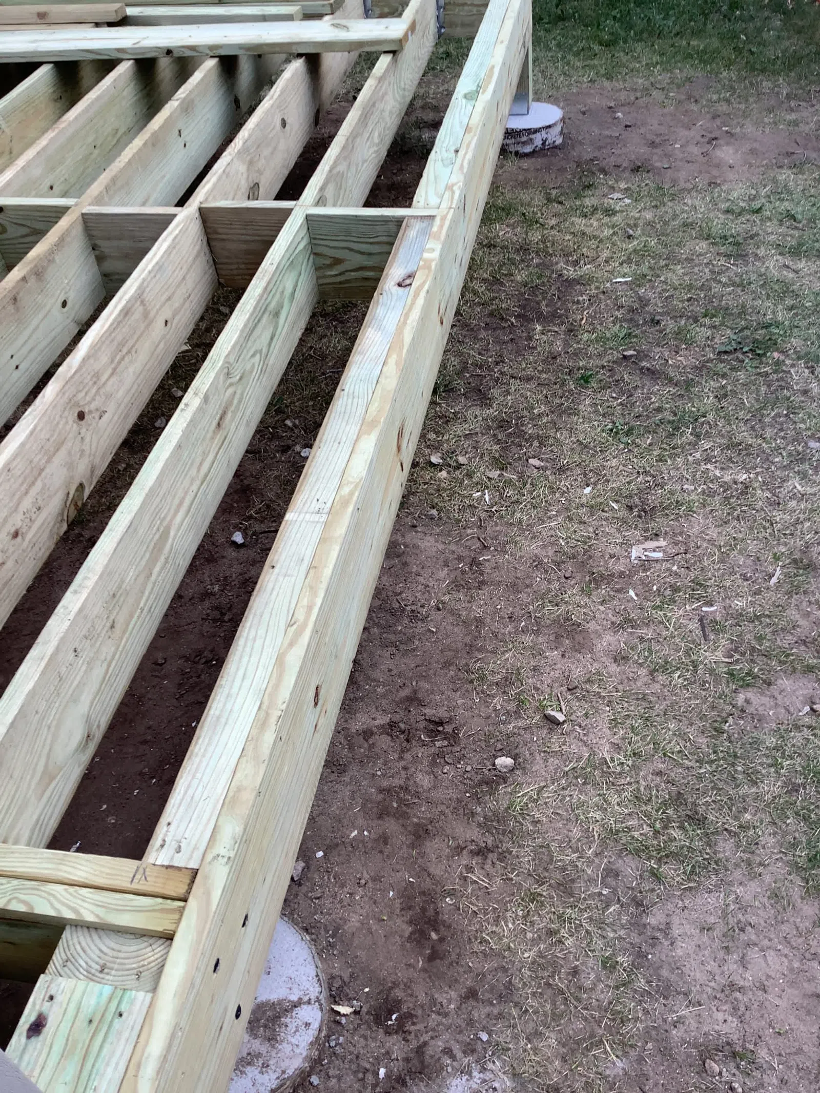 Wooden deck frame under construction on a soil and grass surface with concrete footings.