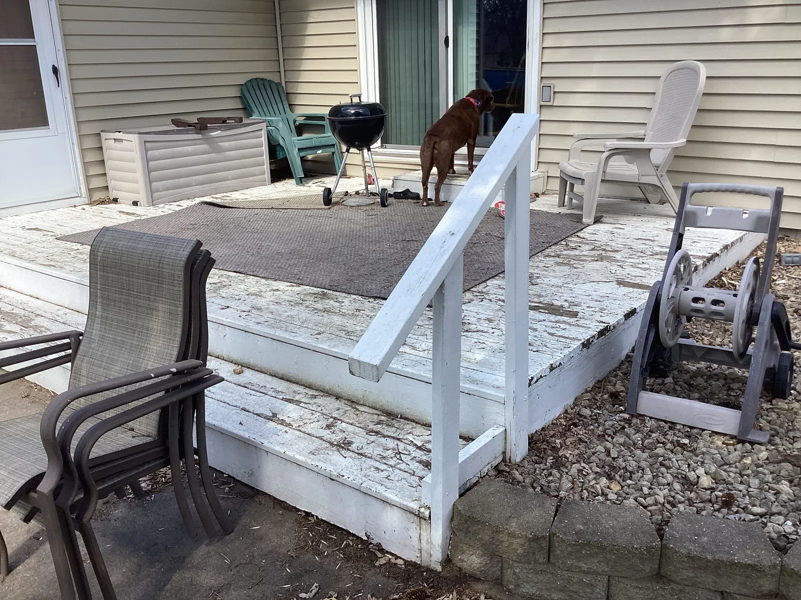 Outdoor wooden deck with peeling white paint, patio chairs, a grill, a dog looking inside sliding glass door.
