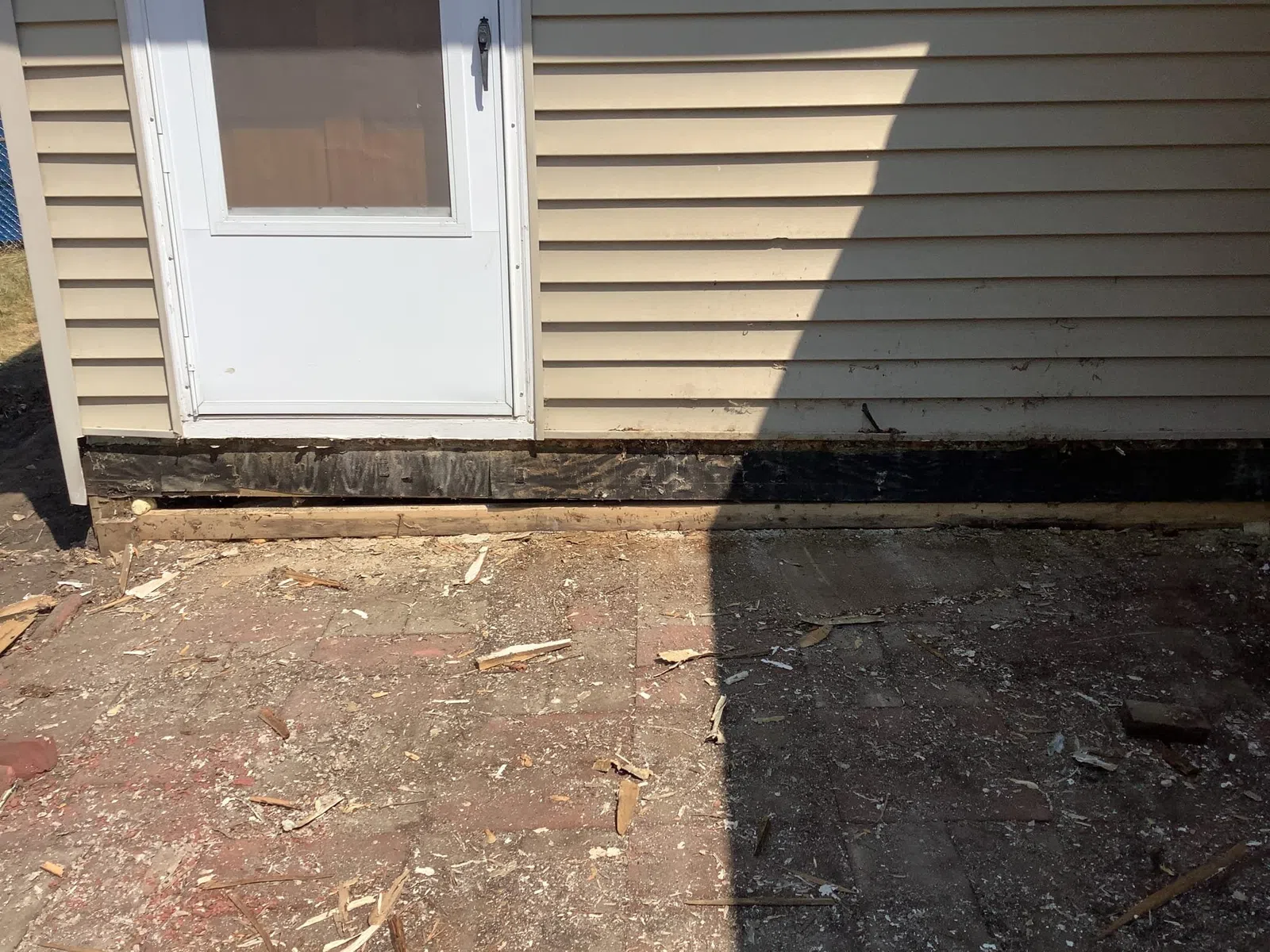 Sunlit area in front of a house with beige siding, a white door, and a brick ground partially covered in debris.