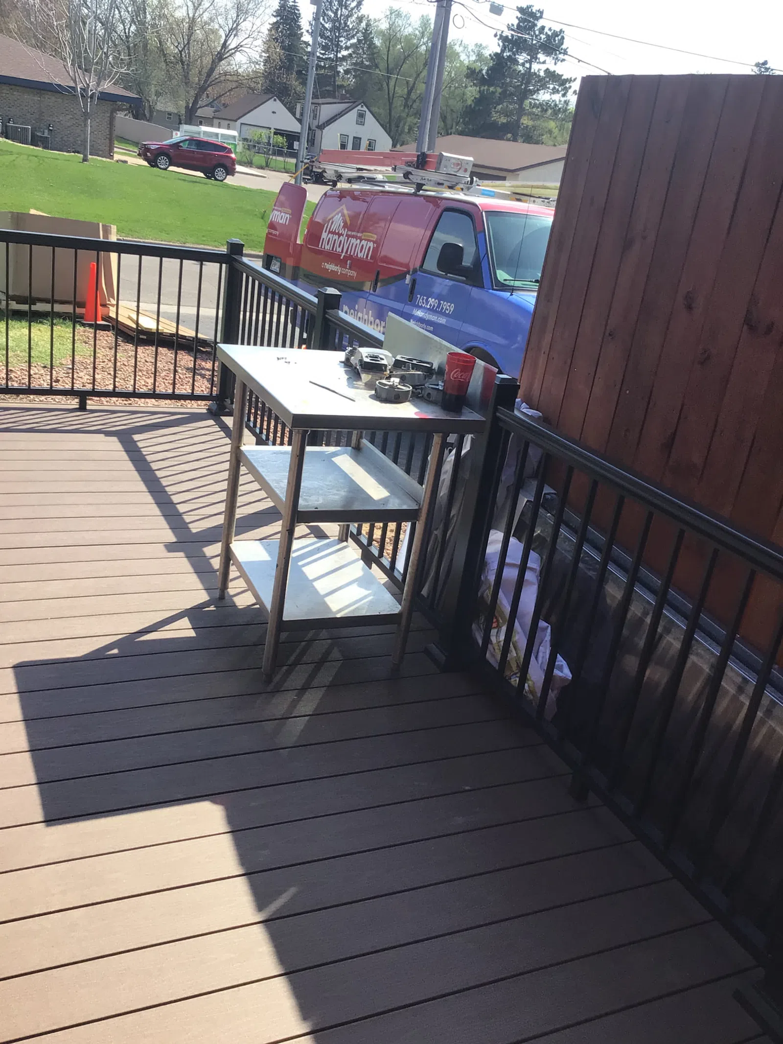 Outdoor deck with metal work table, tools, a Coca-Cola cup, and a handyman service van parked nearby on a sunny day.