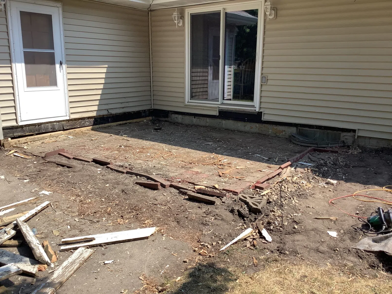 Backyard area with old brick patio partially removed near beige siding house and sliding glass door