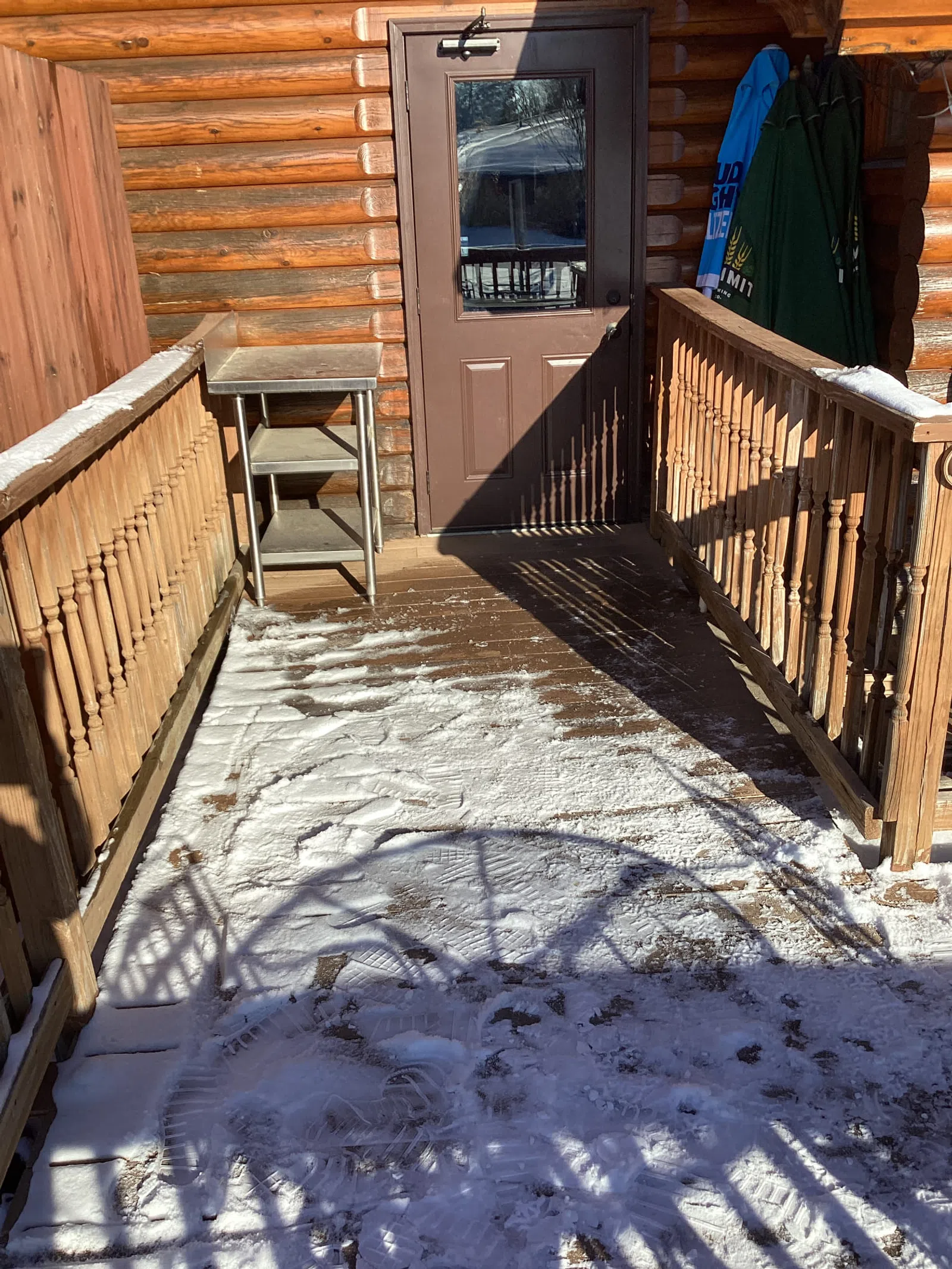 Snow-covered wooden porch with boot prints, railing shadows, and a closed brown door on a log cabin.