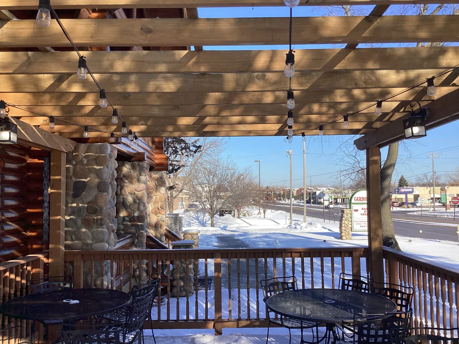 Snow-covered outdoor patio with wooden pergola, string lights, and black metal tables and chairs in a commercial area.
