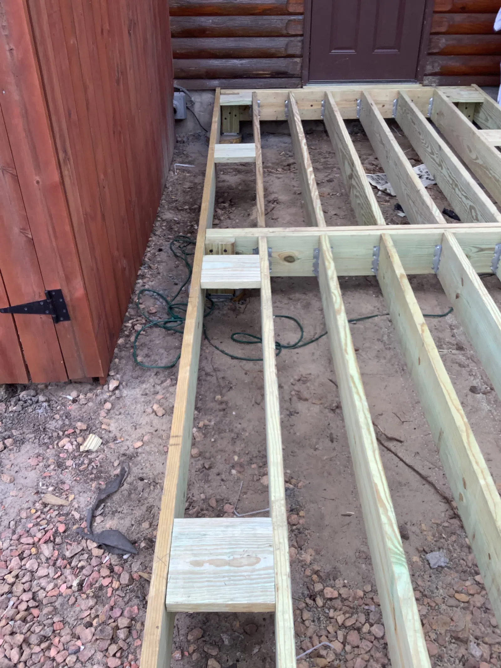 Wooden deck frame construction next to a red shed and log cabin wall on rocky soil ground.