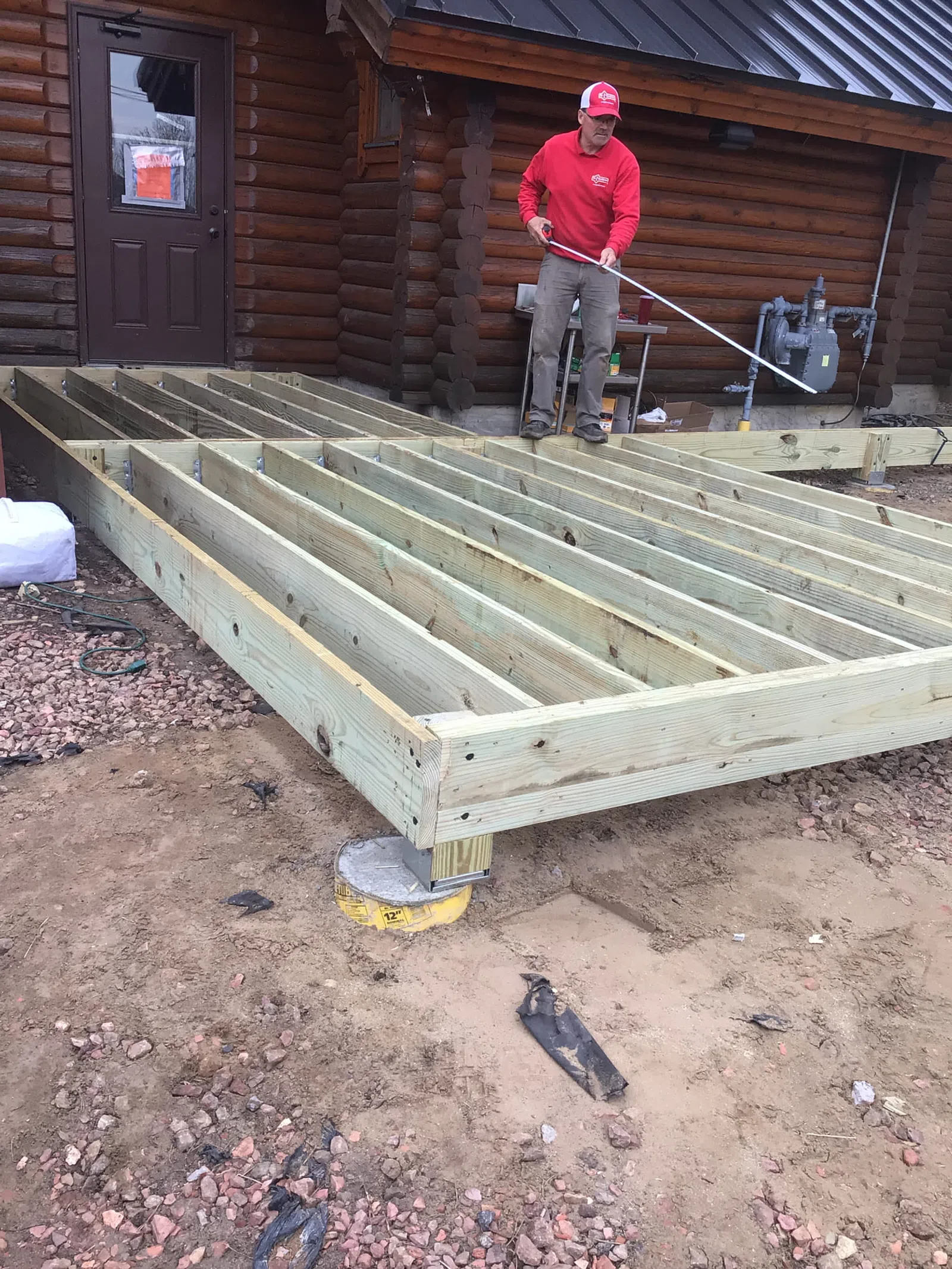 Construction worker building wooden deck frame outside a log cabin on a dirt and gravel surface.