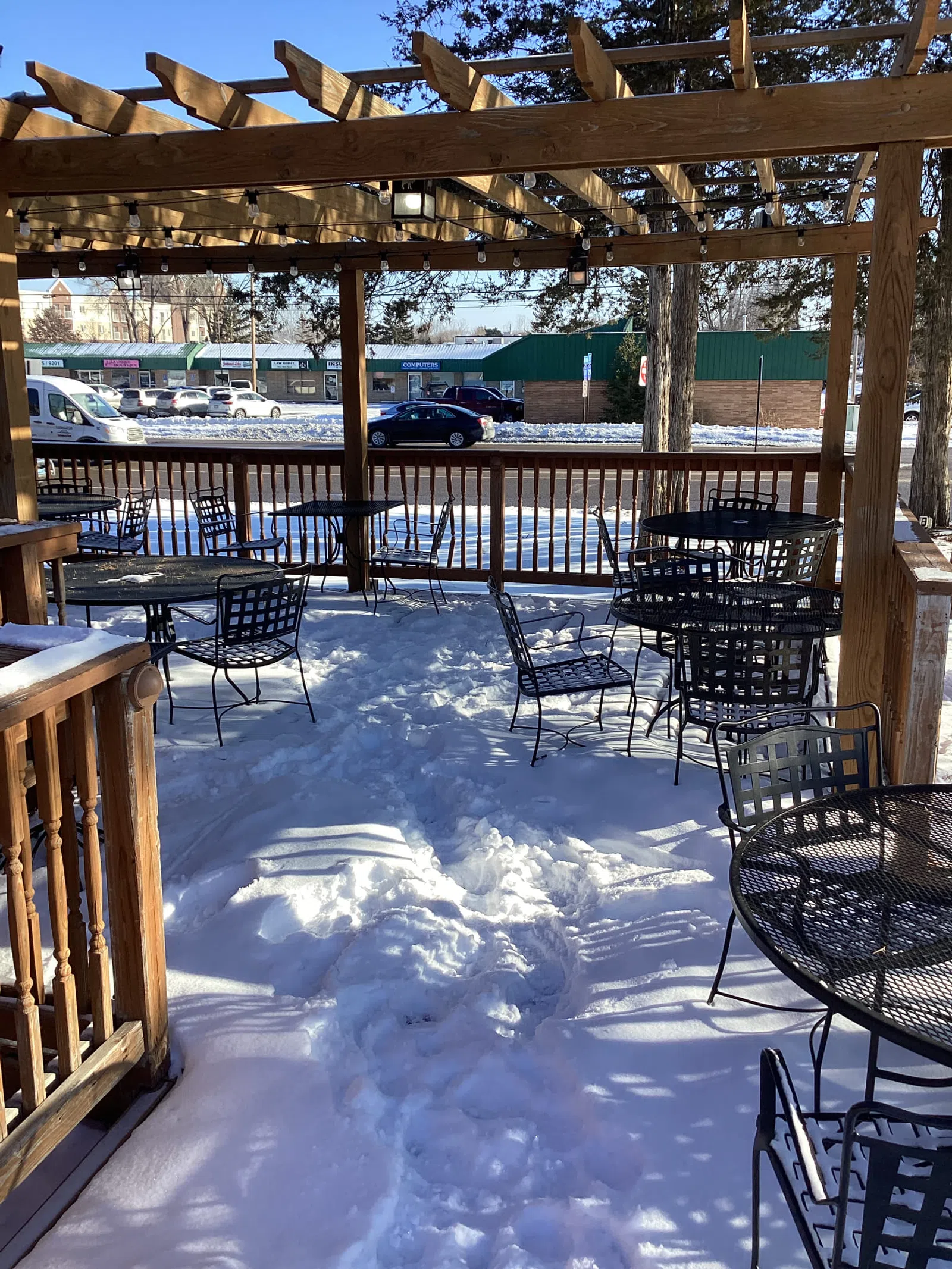 Snow-covered outdoor patio with metal tables and chairs under a wooden pergola on a sunny winter day.