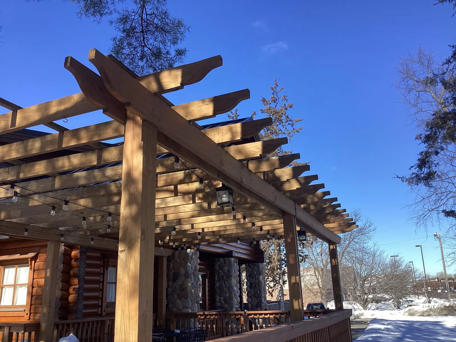 Wooden pergola with string lights on a porch of a log cabin house under clear blue sky in winter.