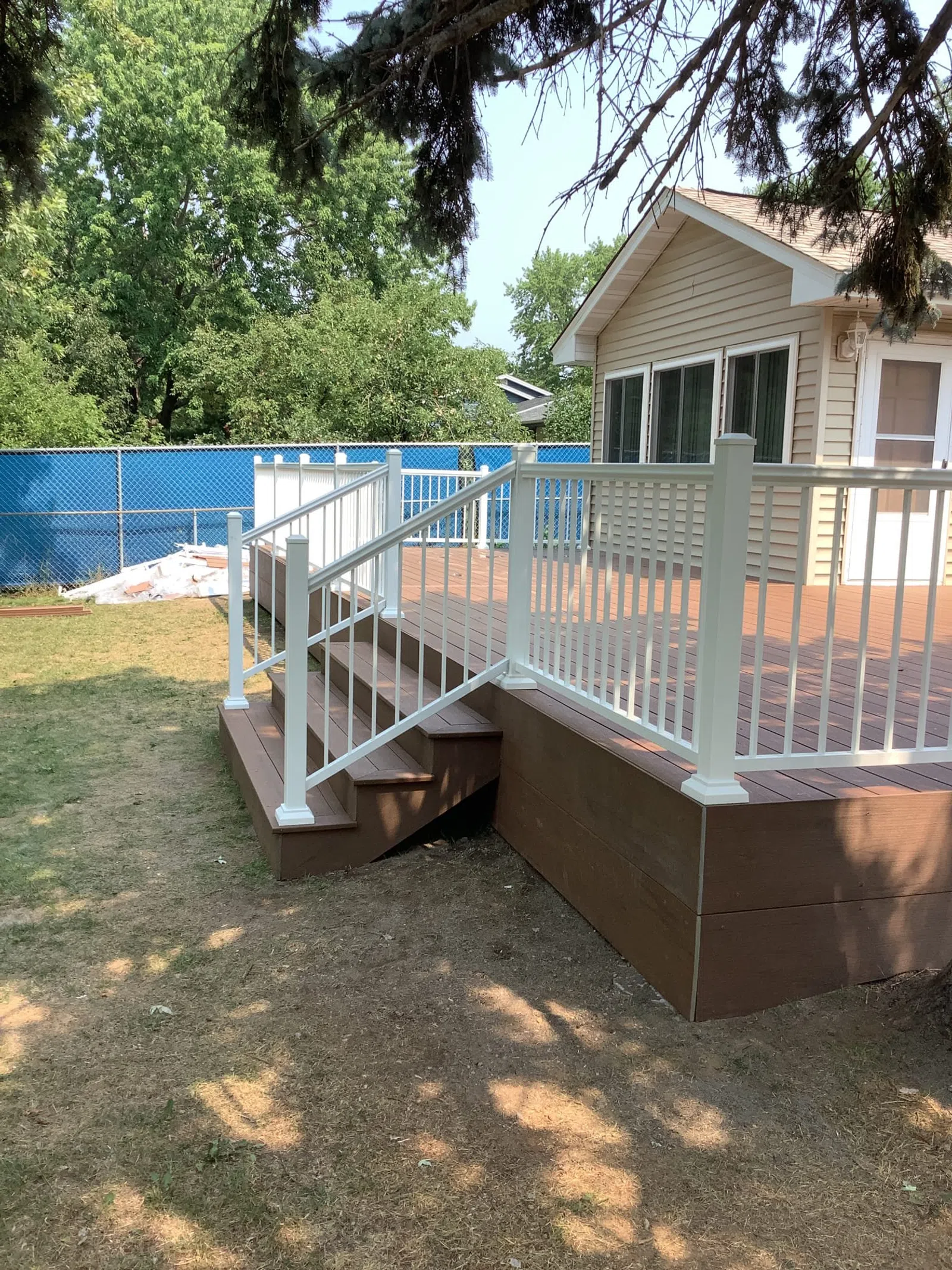 Backyard wooden deck with white railing and steps attached to beige house under trees.