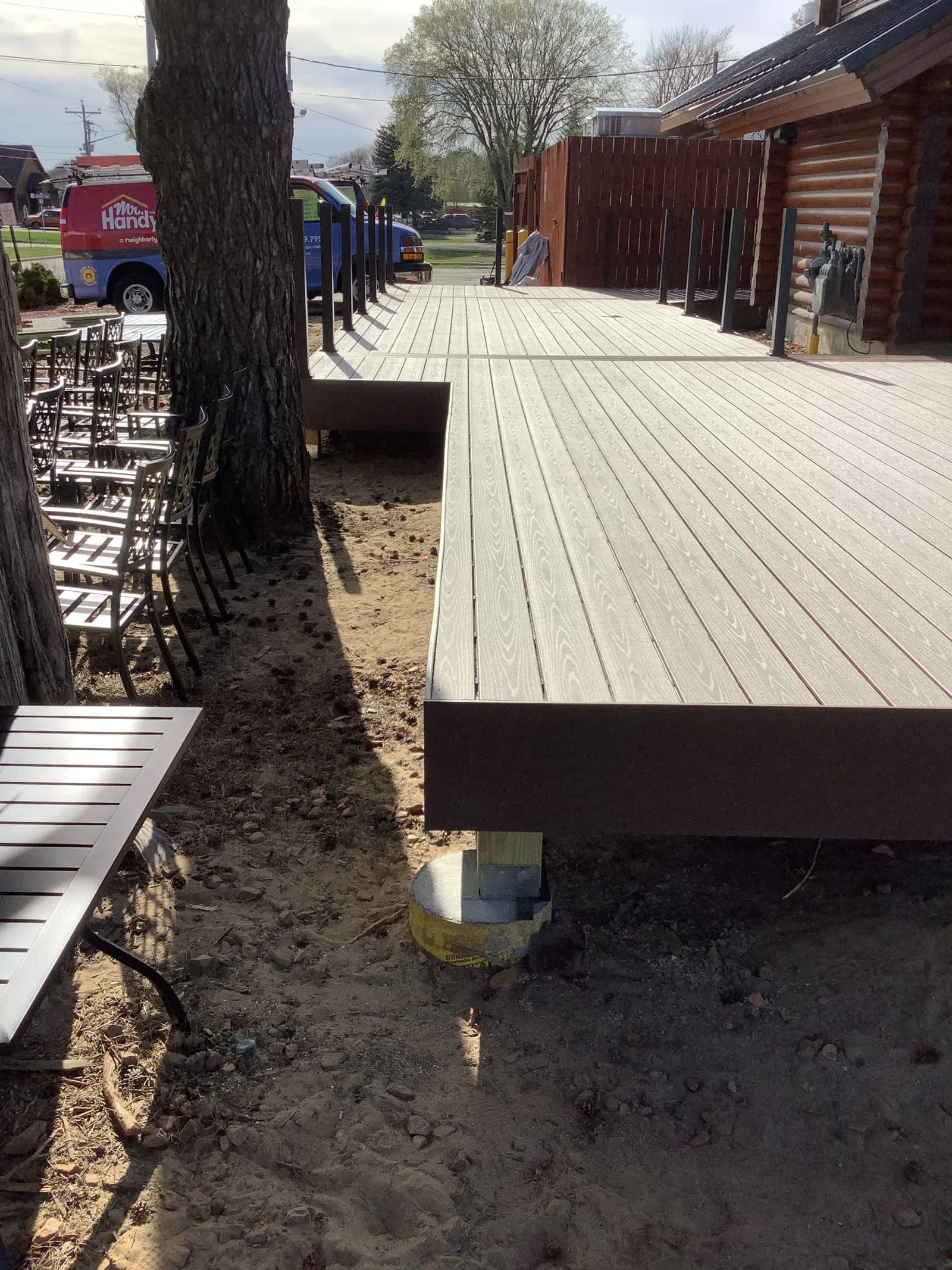 Newly constructed wooden deck beside a log cabin with chairs and a large tree nearby on a sunny day.