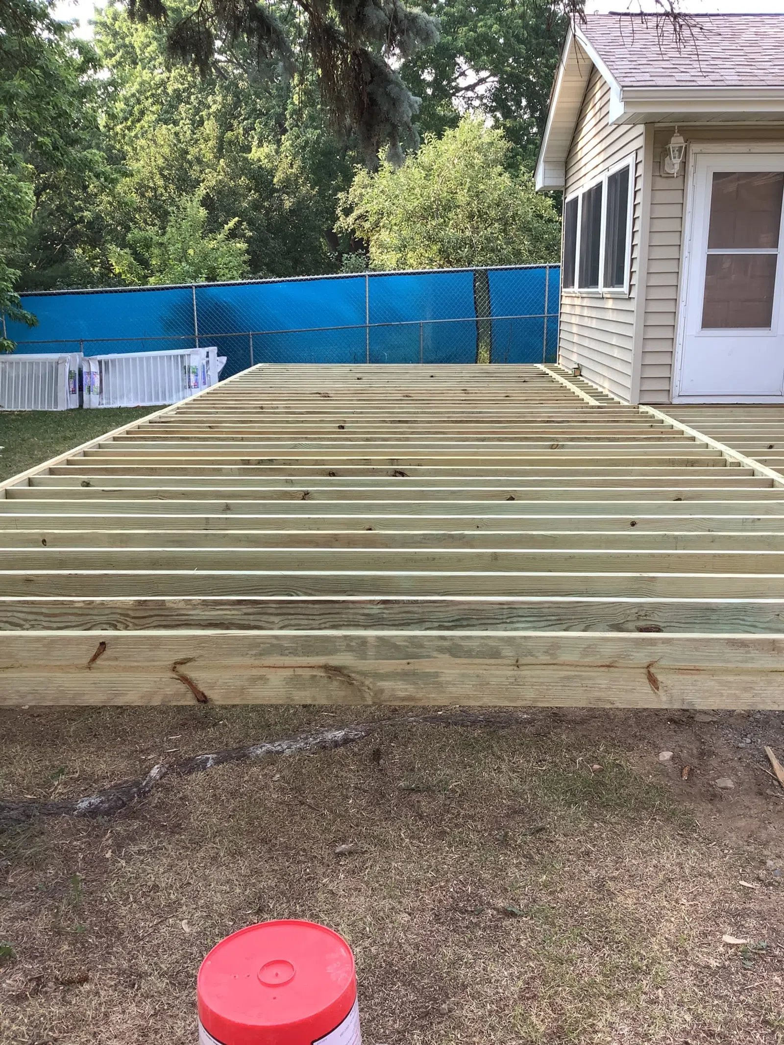 Construction of a wooden deck frame attached to a house in a backyard with trees and a blue fence.