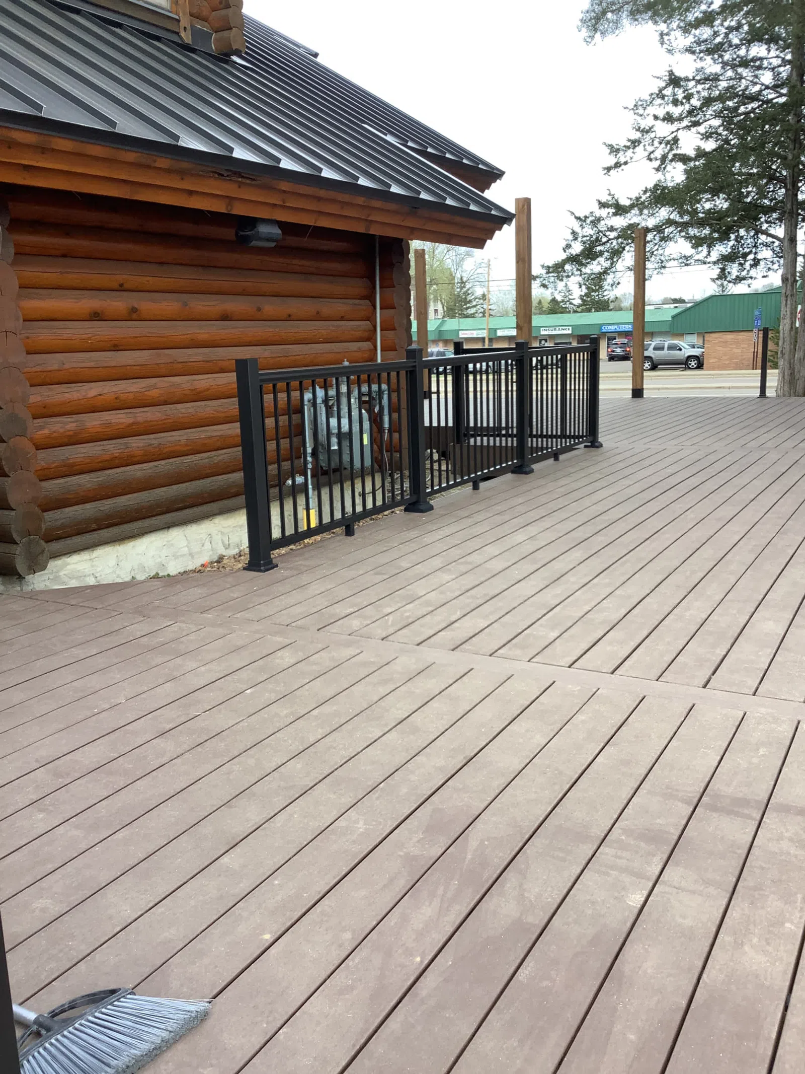 Brown wooden deck next to log cabin with black metal railing and broom on the ground in daytime.