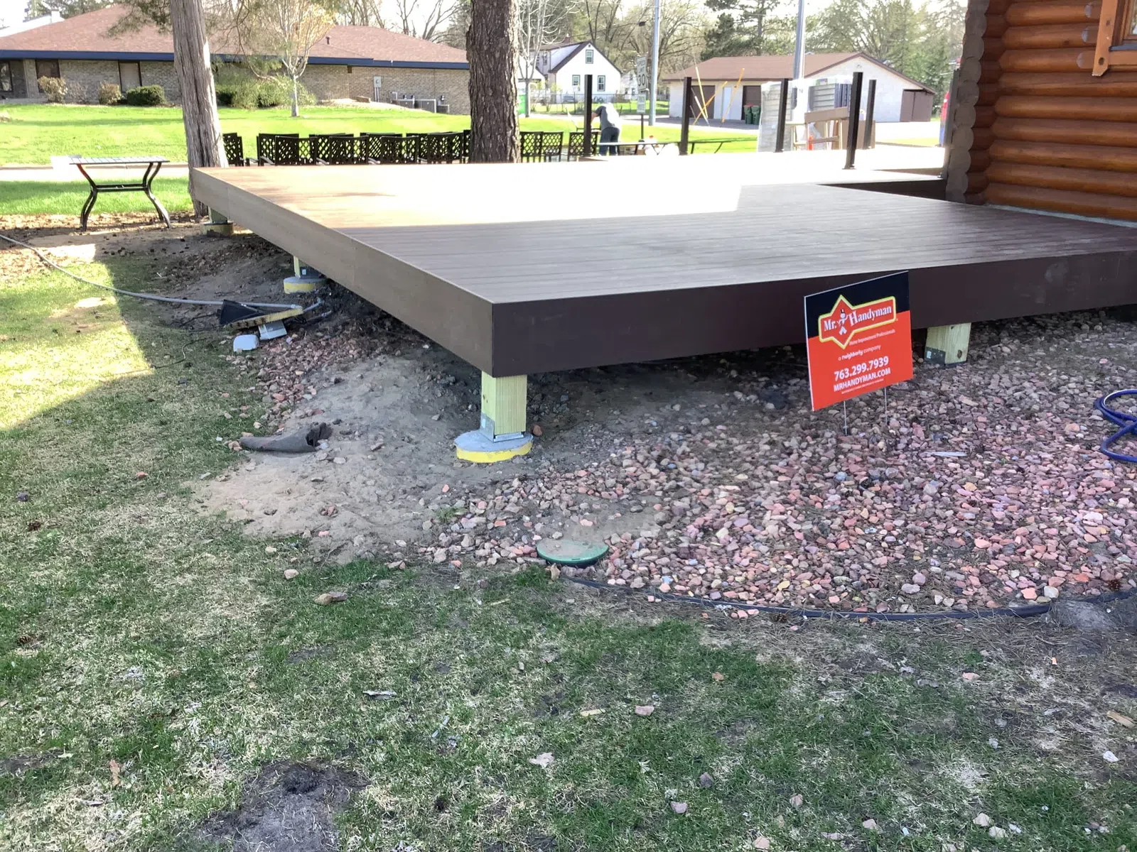 Raised wooden deck platform supported by posts over gravel and grass near a log cabin exterior.