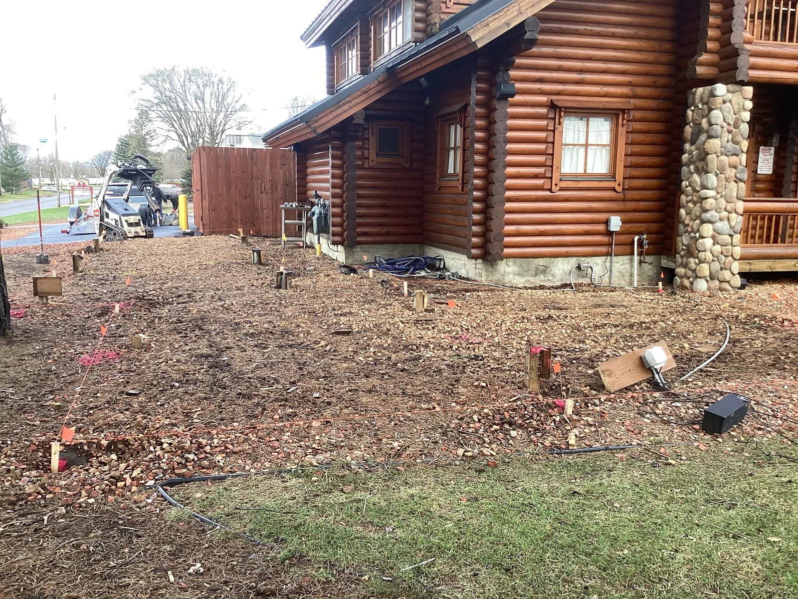 Prepared ground with stakes and markings next to a log cabin under construction with stone pillar details.